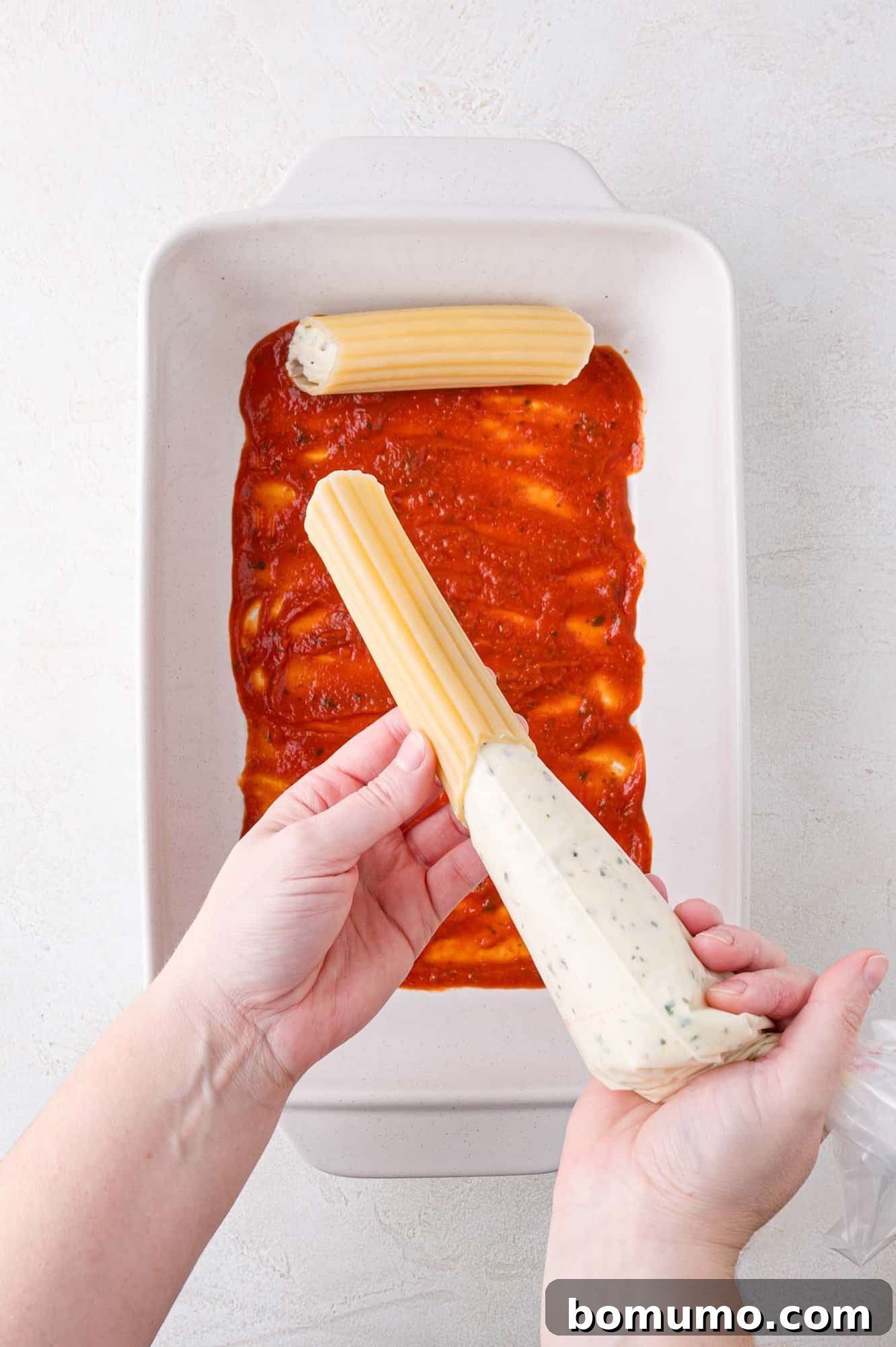 Hands fill cannelloni with ricotta mixture using a piping bag over a baking dish containing tomato sauce.