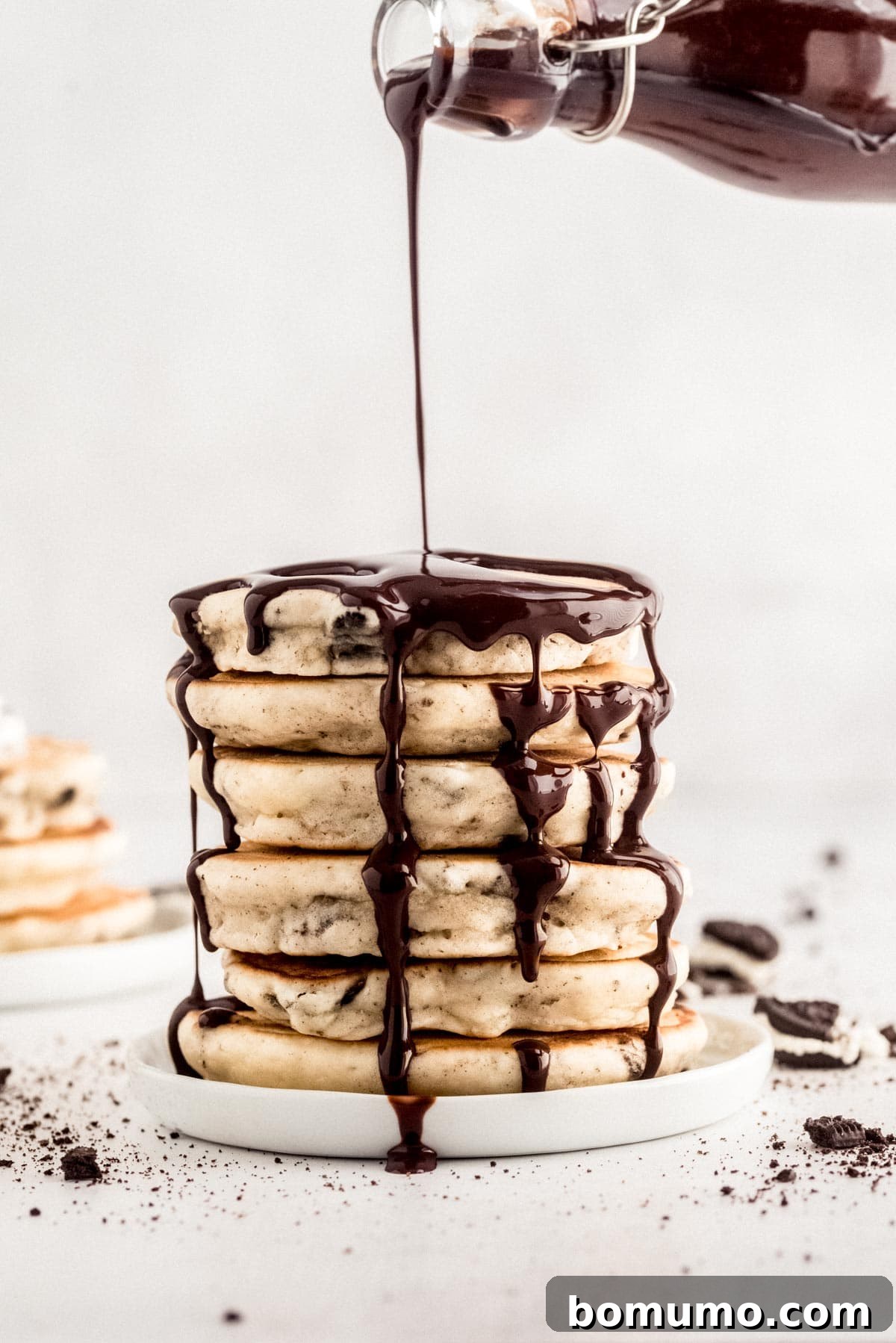 A beautifully presented stack of fluffy pancakes generously drizzled with rich chocolate syrup on a pristine white plate, surrounded by a scattering of crumbs, inviting a delicious breakfast.