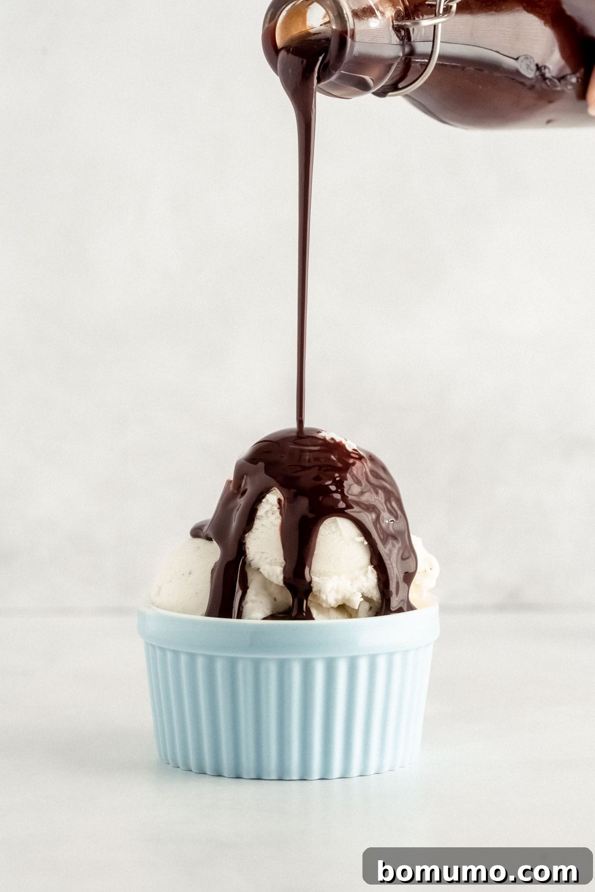 Rich, dark chocolate syrup being generously poured over a scoop of creamy vanilla ice cream nestled in a light blue bowl, set against a clean background.