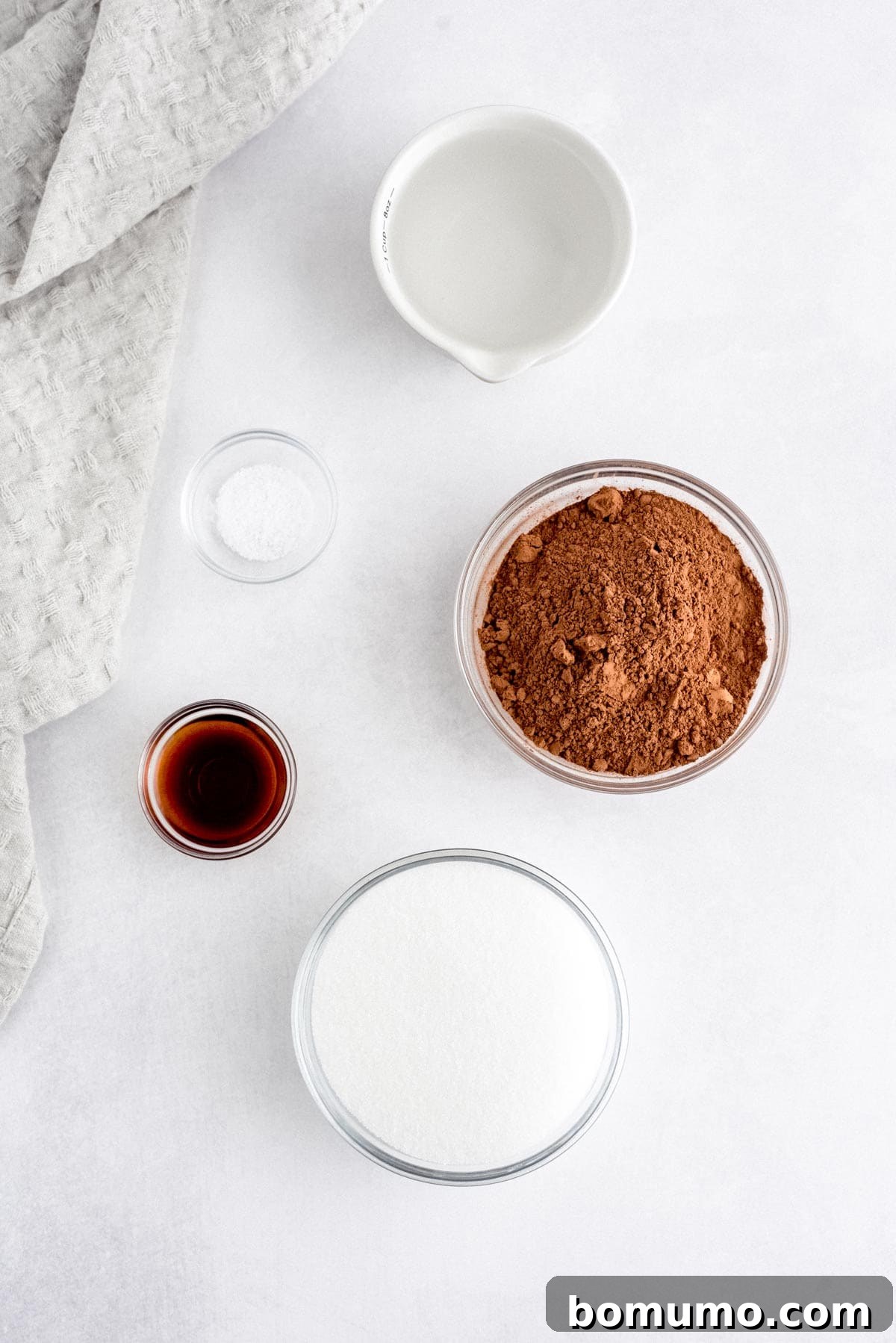 Neatly arranged bowls containing the essential ingredients for chocolate syrup: cocoa powder, granulated sugar, vanilla extract, salt, and water, displayed on a clean white surface with a textured gray cloth.