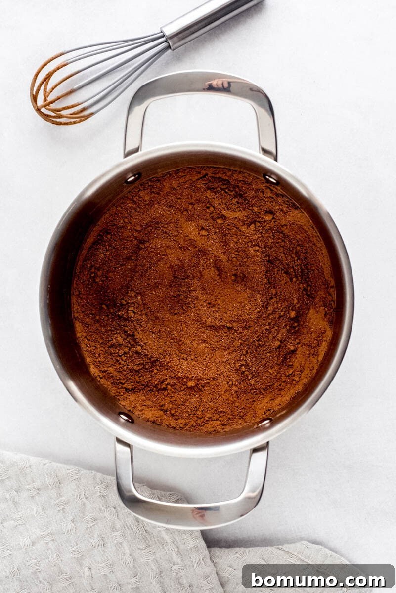 A pot filled with a well-mixed blend of cocoa powder, sugar, and salt on a light surface, with a whisk resting nearby, showing the uniform dry mixture.