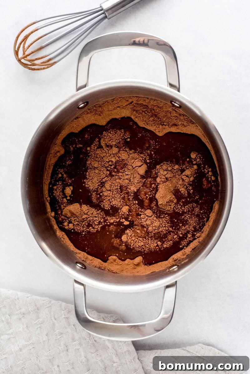 A stainless steel pot on a surface, containing the combined wet and dry chocolate mixture, with a whisk and a cloth nearby, illustrating the initial stage of heating.