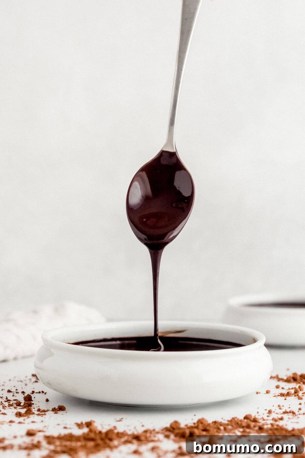 A spoon drizzles lustrous chocolate sauce into a small, round white dish, with a light dusting of cocoa powder scattered on a white surface, creating an elegant presentation.