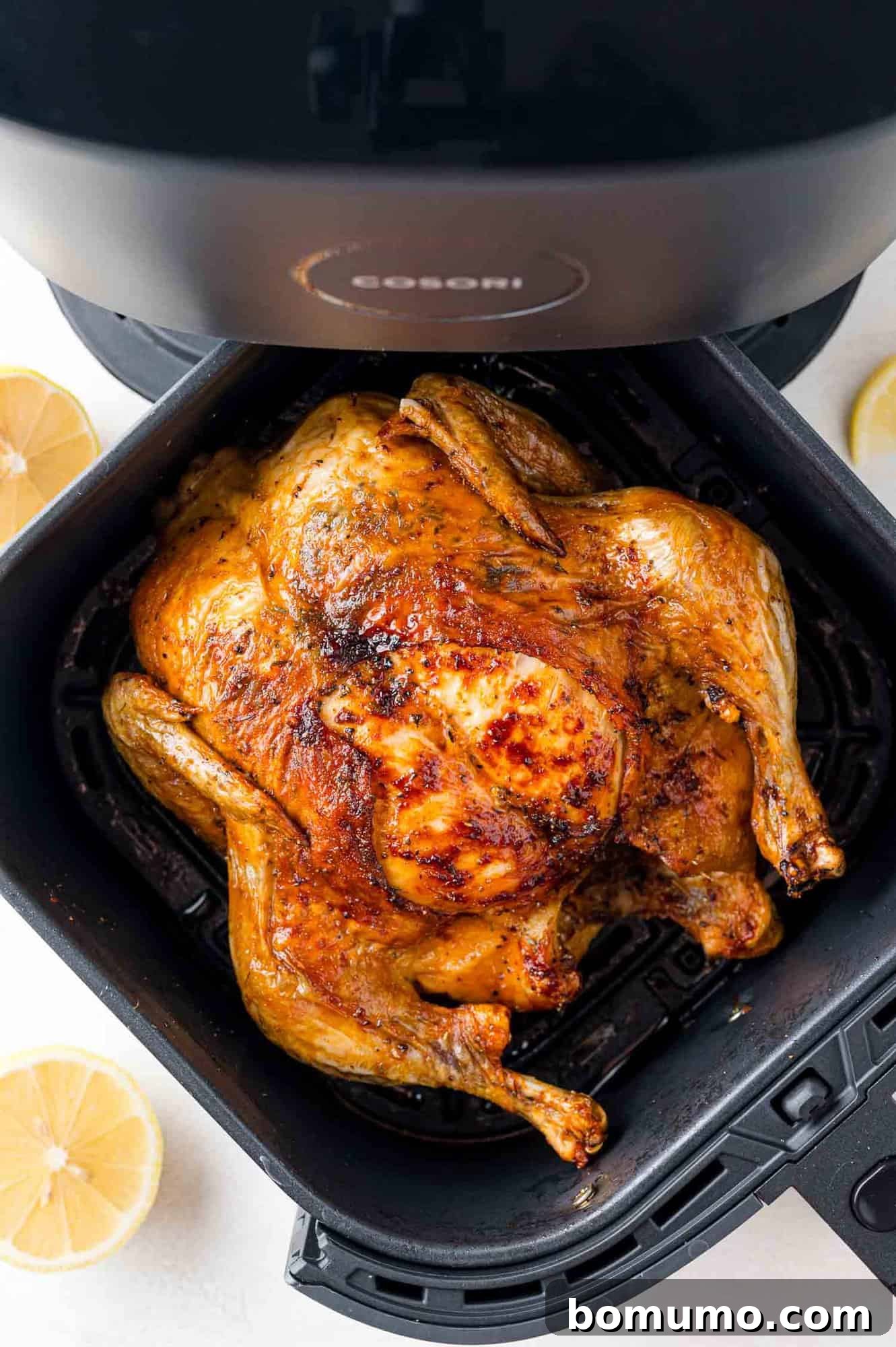 Roasted whole chicken with golden brown skin in an air fryer basket, surrounded by lemon slices.