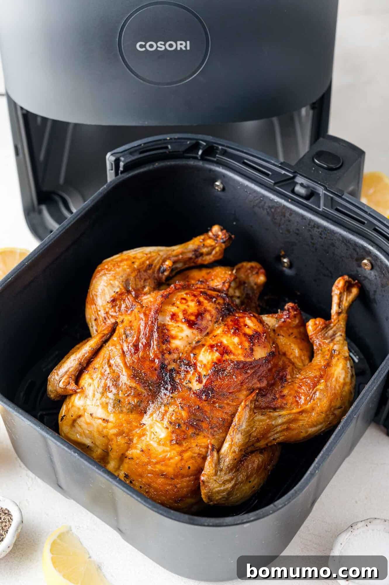 Cooked whole chicken in a black air fryer basket, with the air fryer visible in the background. Lemon slices and seasoning are nearby.