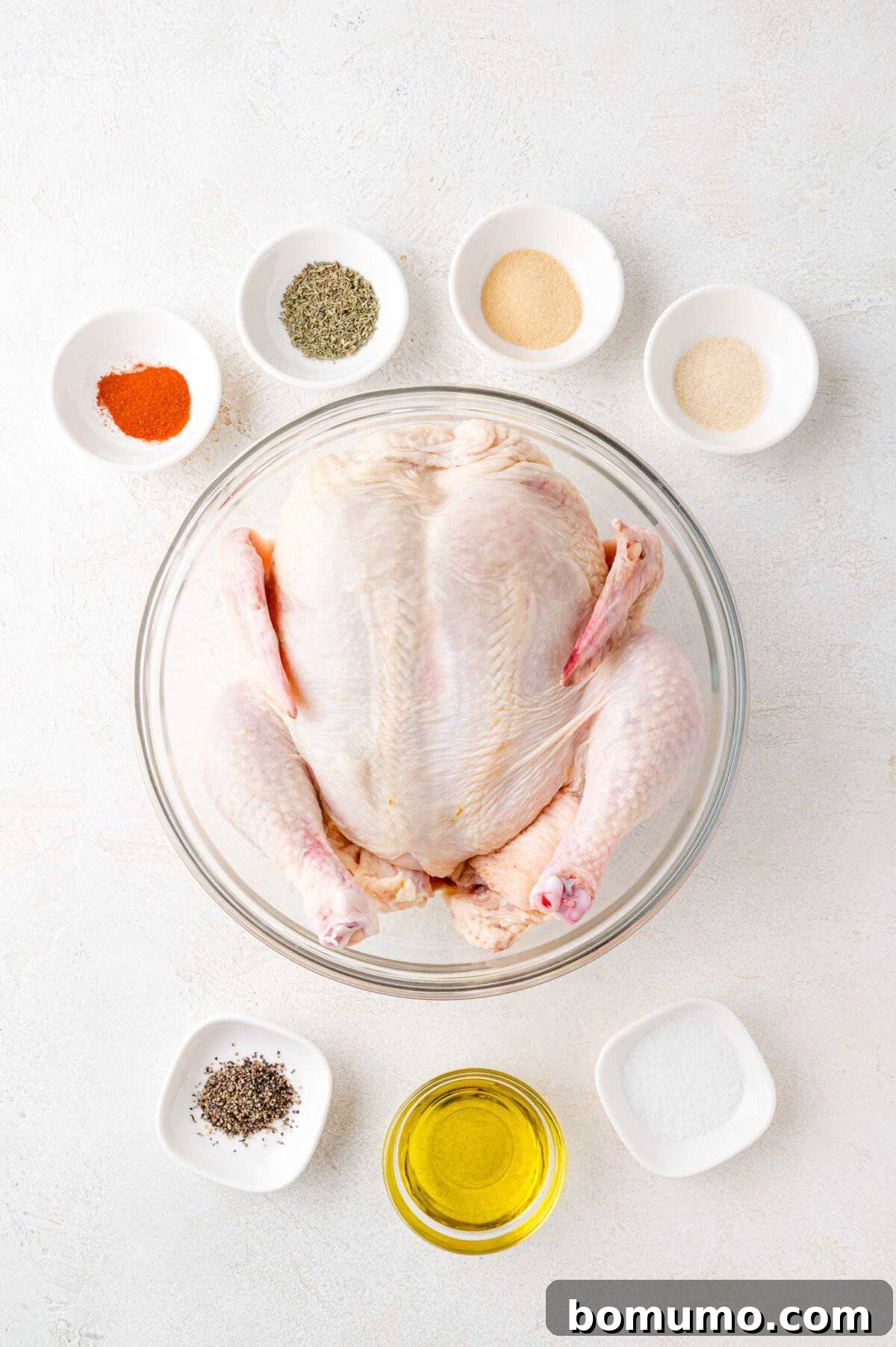 A raw whole chicken in a glass bowl surrounded by small bowls of spices, including black pepper, salt, garlic powder, and oil, on a white surface.