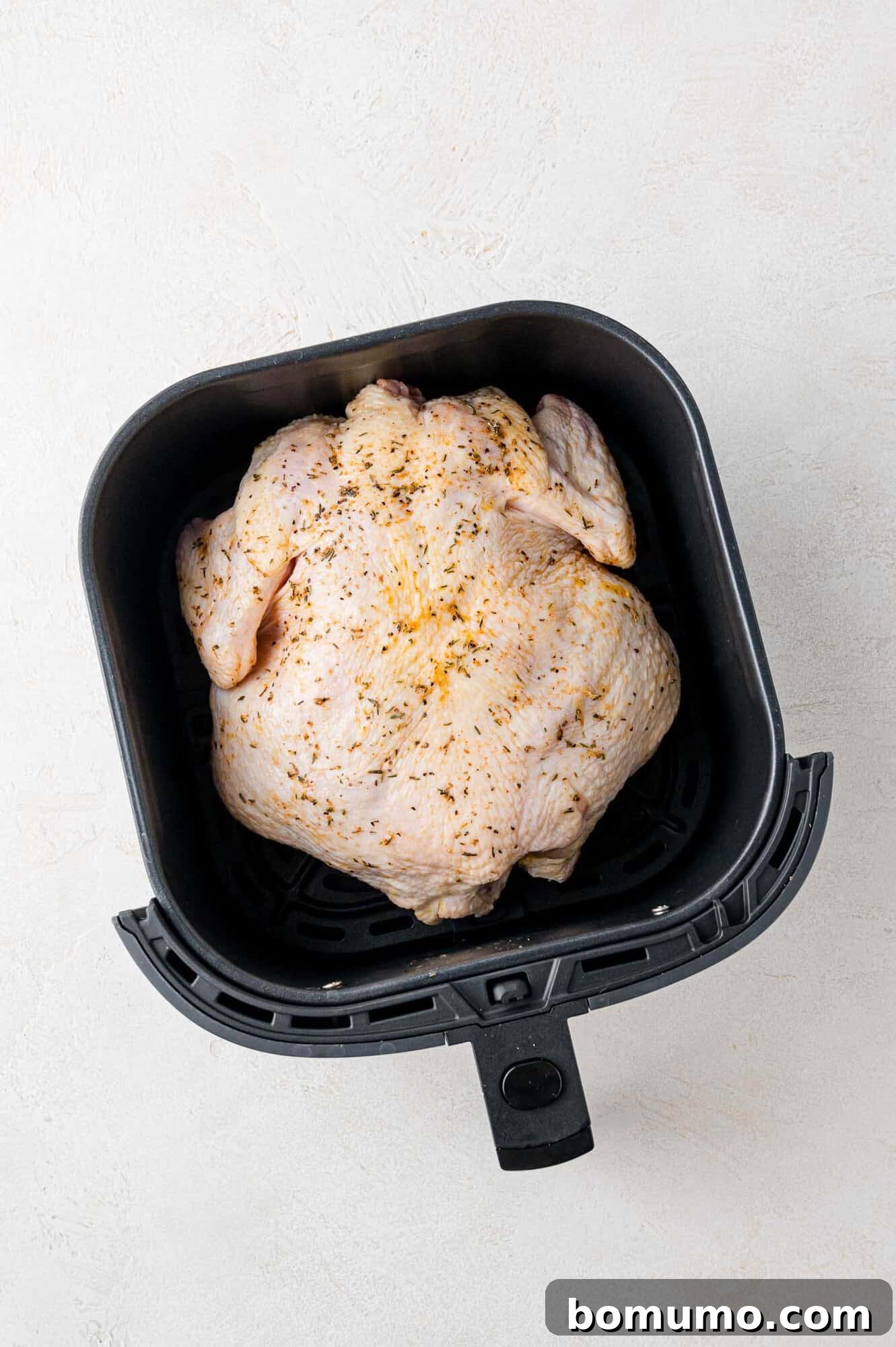 Whole raw chicken seasoned with spices placed inside a black air fryer basket on a light background.