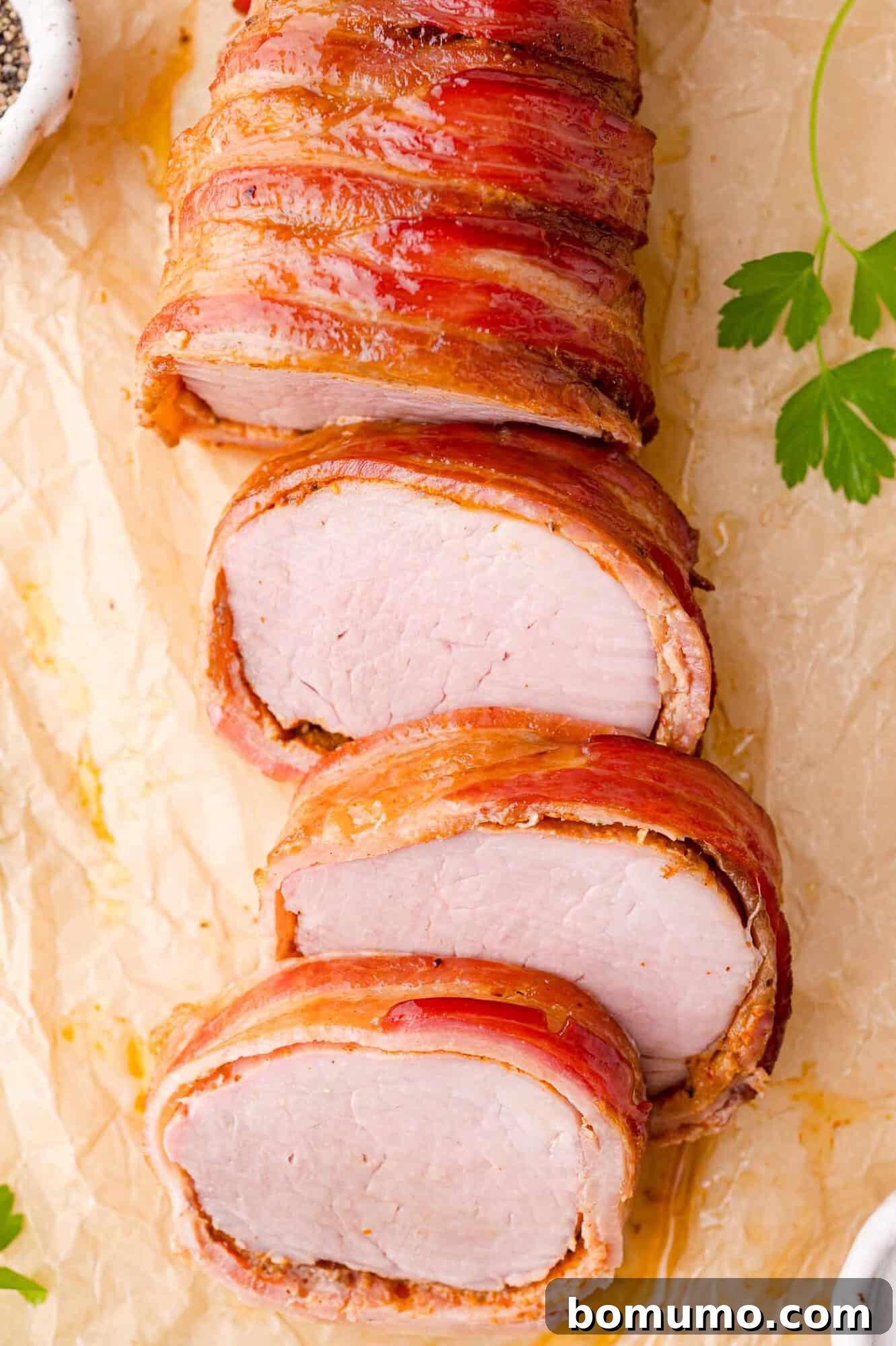 Sliced bacon-wrapped pork tenderloin on parchment paper, garnished with parsley.