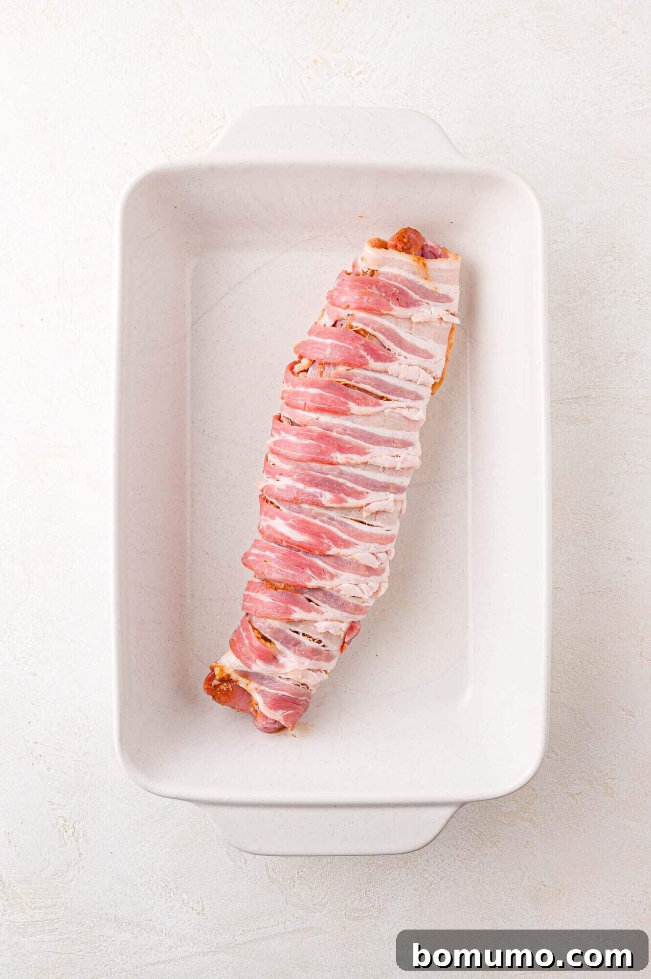 A piece of bacon-wrapped pork tenderloin on a white baking dish.