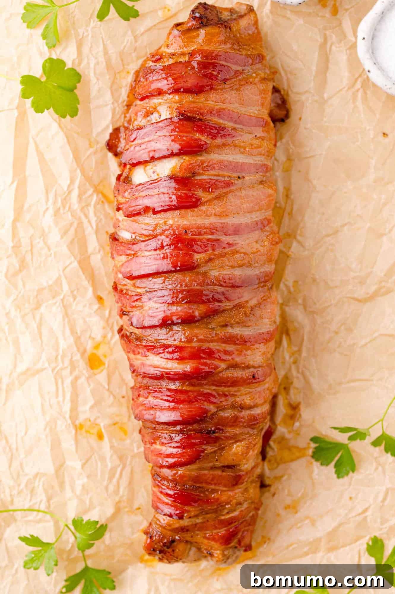 A bacon-wrapped pork tenderloin on parchment paper, garnished with parsley leaves.