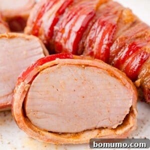 Sliced bacon-wrapped pork tenderloin on a white plate, showing a juicy interior and crispy bacon exterior.