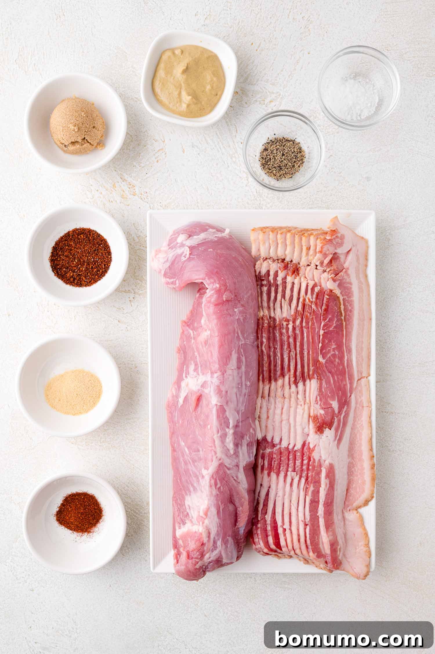 Raw pork tenderloin and bacon on a white tray, surrounded by small bowls of spices and seasonings on a light surface.