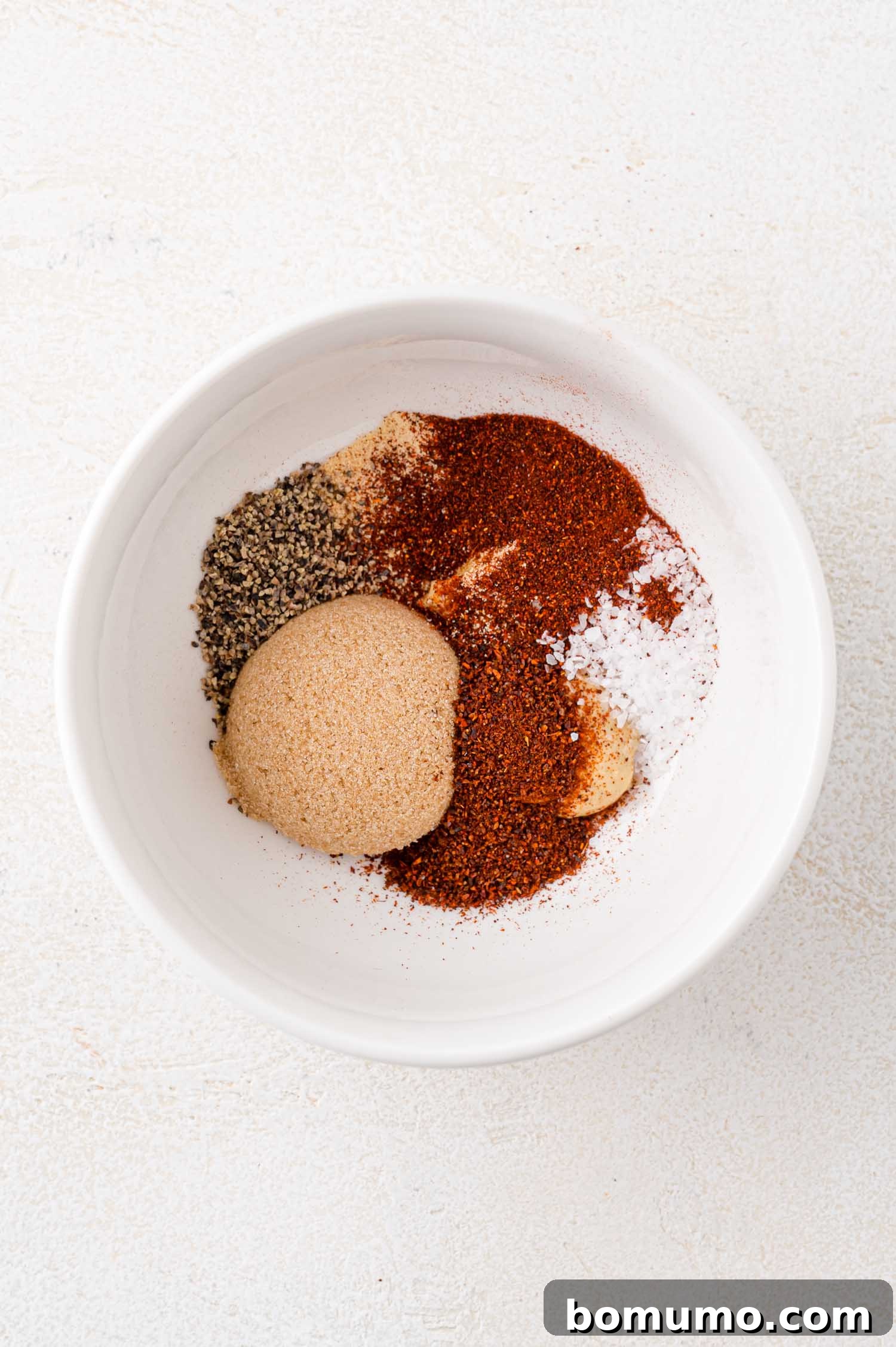 A white bowl filled with various spices, including brown sugar, chili powder, black pepper, and salt, on a light surface.