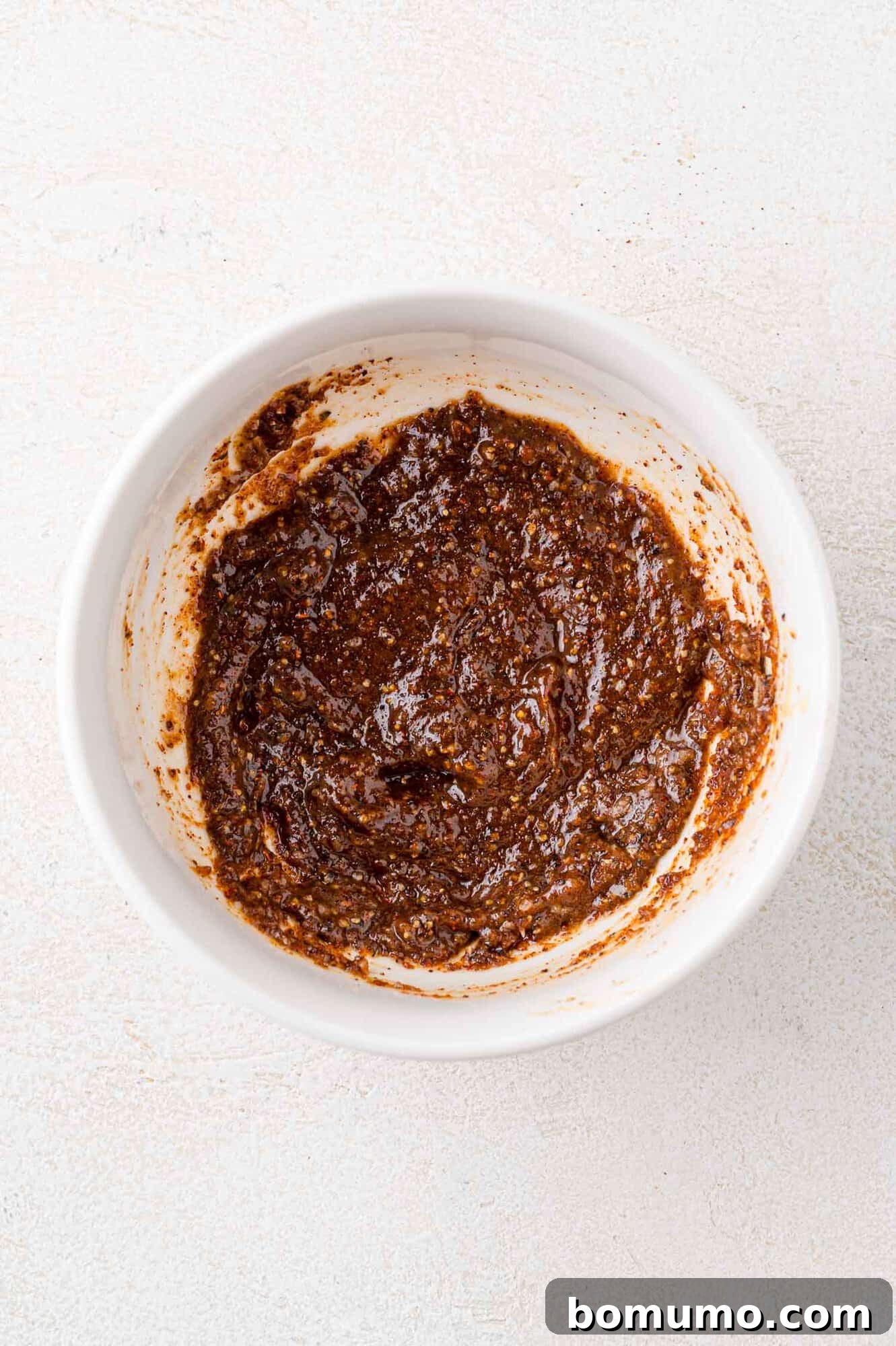 White bowl containing a thick, dark brown paste with a textured surface, on a light background.