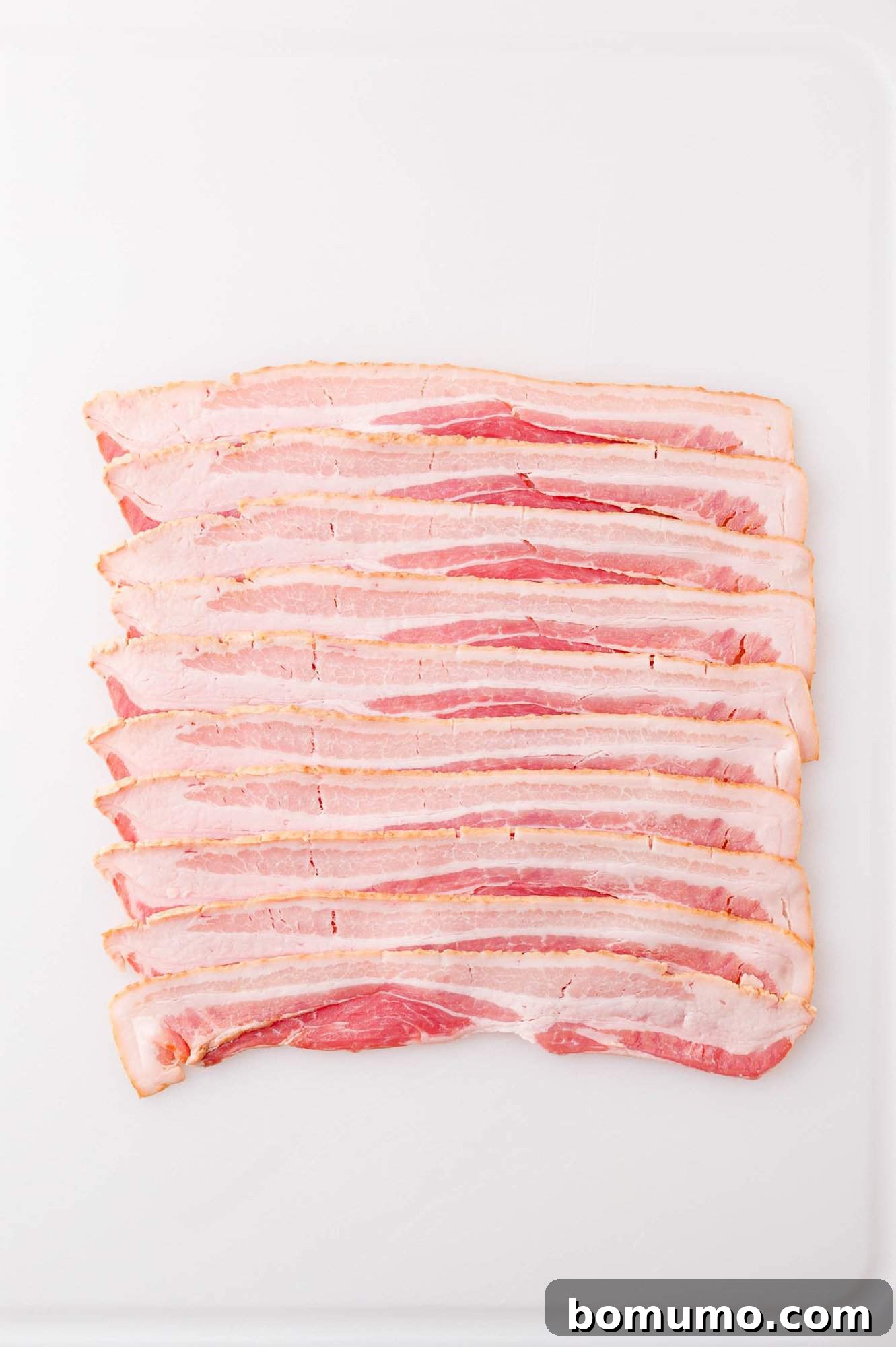 Several uncooked bacon strips are laid out in a neat row on a white surface.