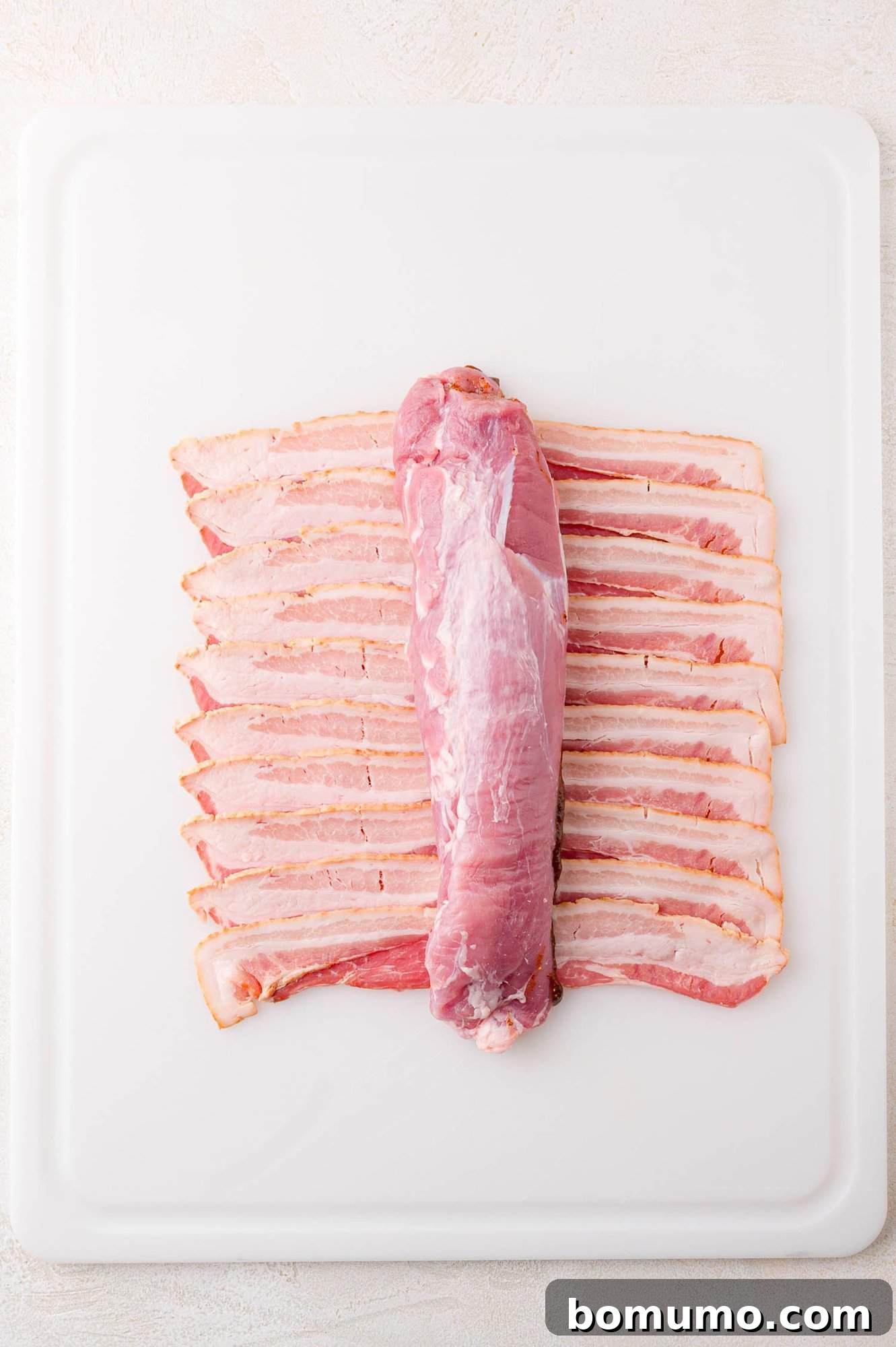 A raw pork tenderloin is placed on top of several strips of uncooked bacon, arranged neatly in rows, on a white cutting board.