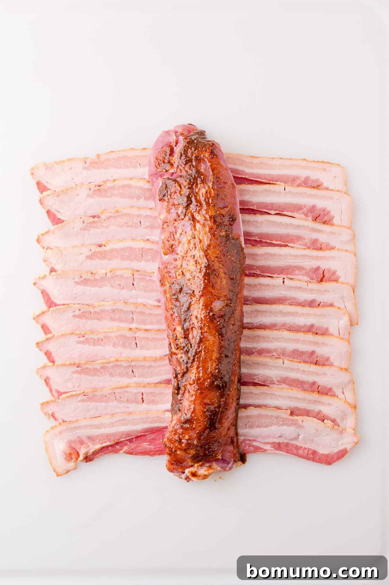 A row of sliced bacon is laid out on a white surface with a seasoned pork tenderloin placed on top.