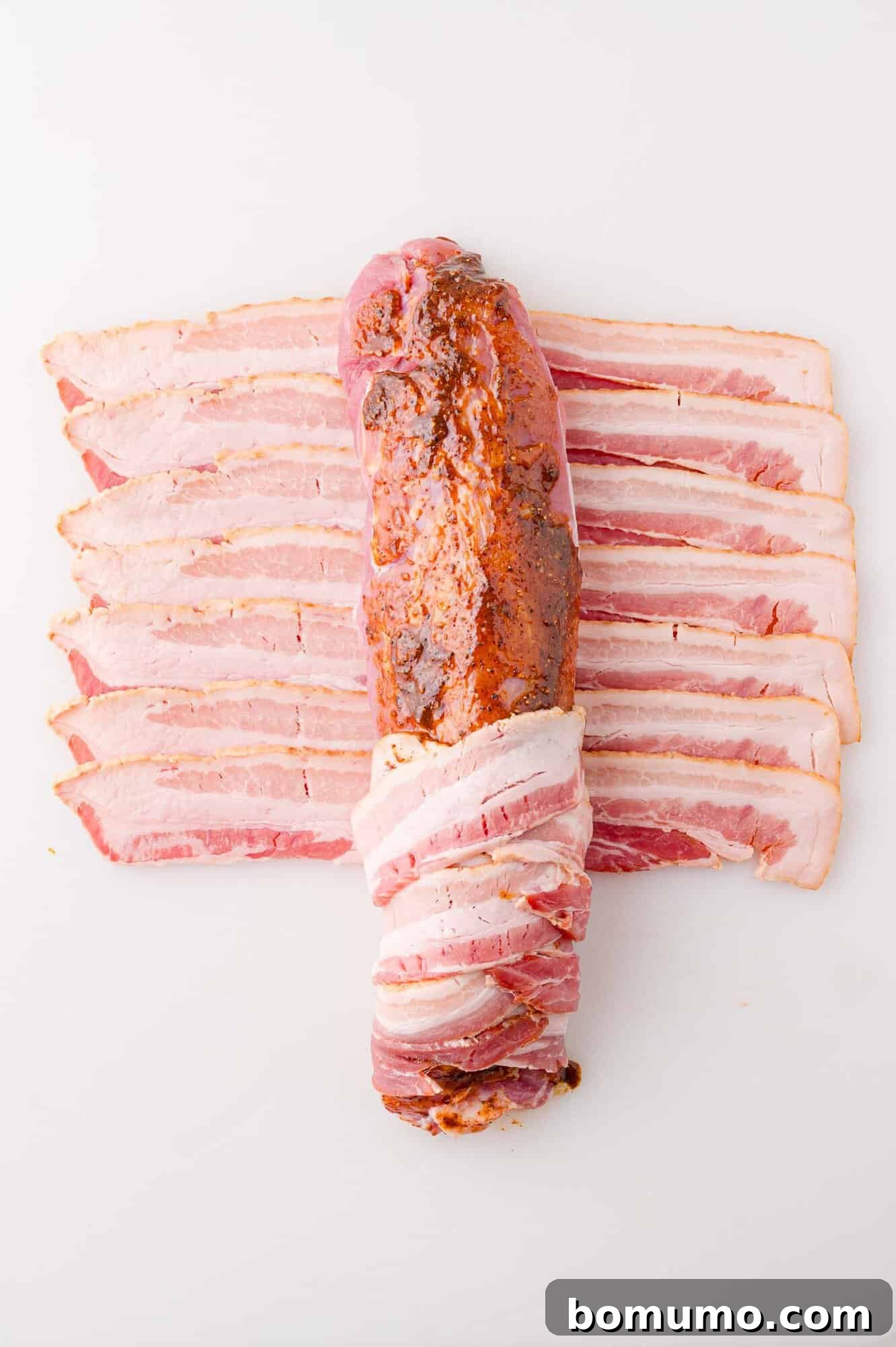 Raw pork tenderloin seasoned and wrapped with strips of uncooked bacon, displayed on a white surface.