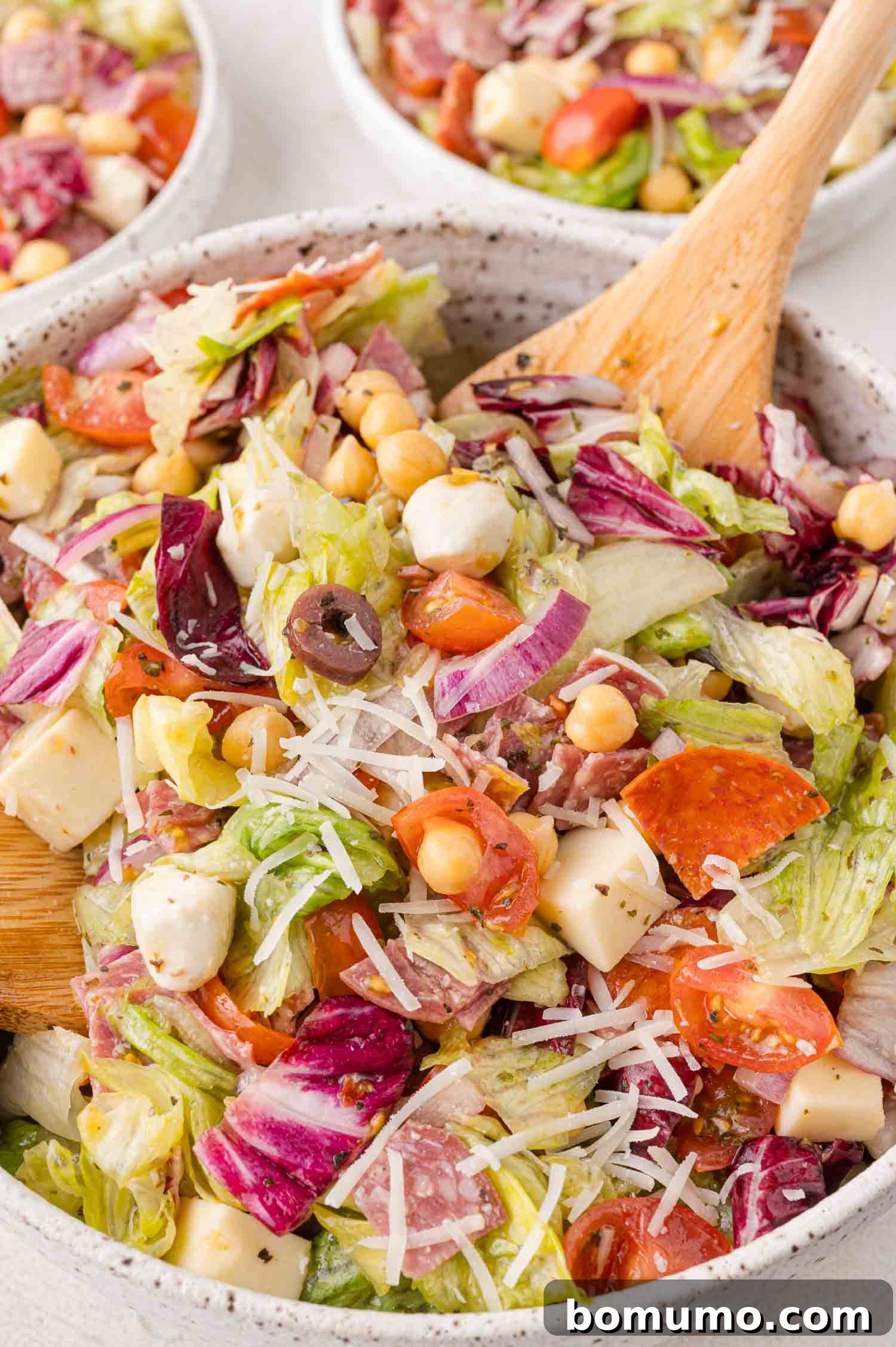 Chopped Salad Perfection 3 A mixed salad in a bowl featuring lettuce, tomatoes, cheese cubes, olives, chickpeas, and grated cheese, with a wooden spoon. Other bowls of salad are visible in the background.