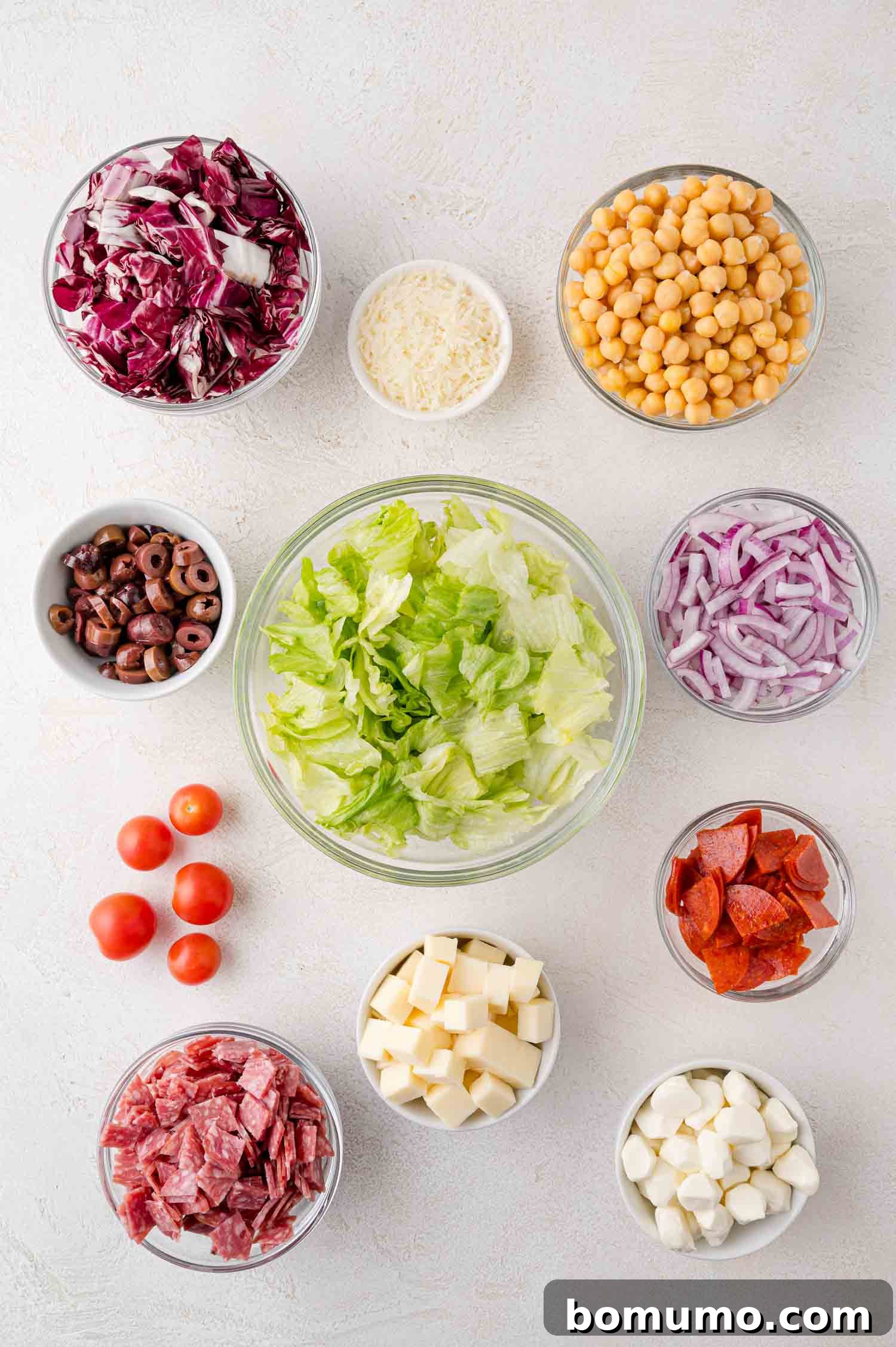 Chopped Salad Perfection 4 Bowls of salad ingredients including lettuce, chickpeas, radicchio, red onions, cheese, cherry tomatoes, olives, pepperoni, and salami, arranged on a white surface.