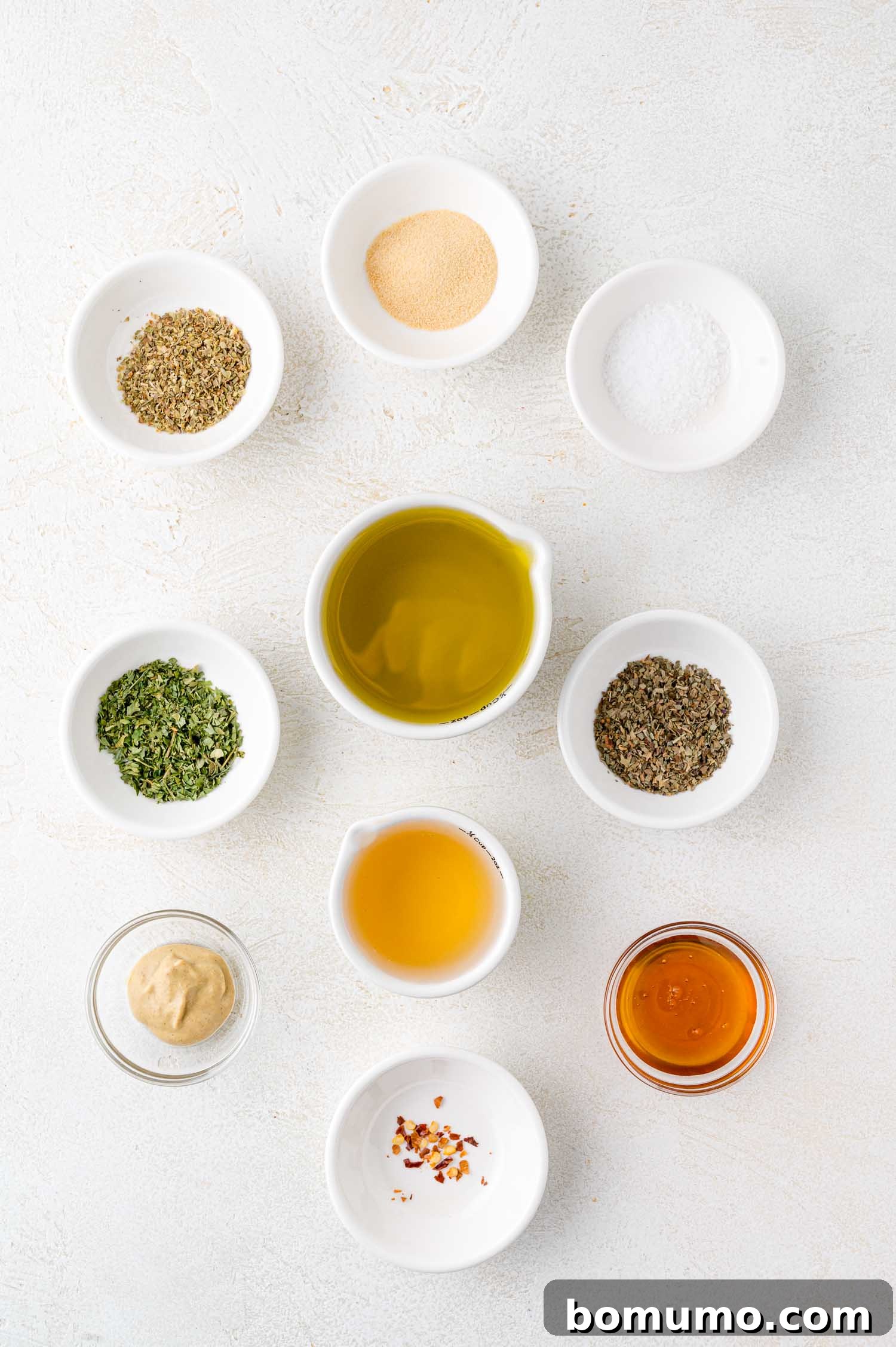 Chopped Salad Perfection 6 An assortment of small white bowls containing spices, herbs, oil, vinegar, mustard, and honey arranged on a white surface.