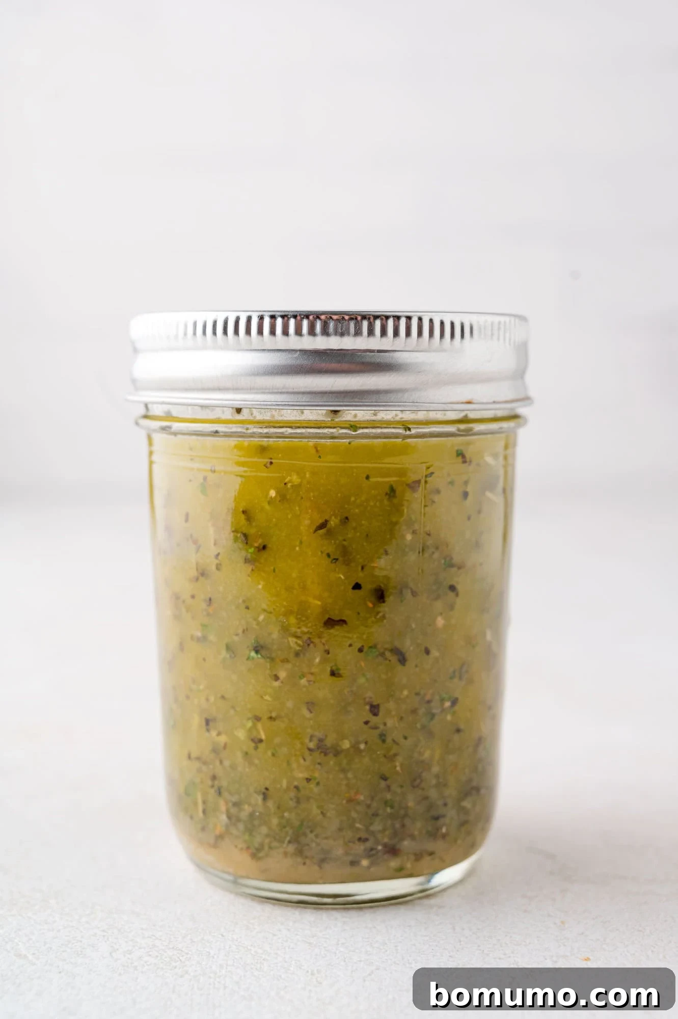 Chopped Salad Perfection 7 A clear, lidded jar filled with a greenish mixture that appears to be a sauce or condiment with visible herb and spice specks, standing on a light background.