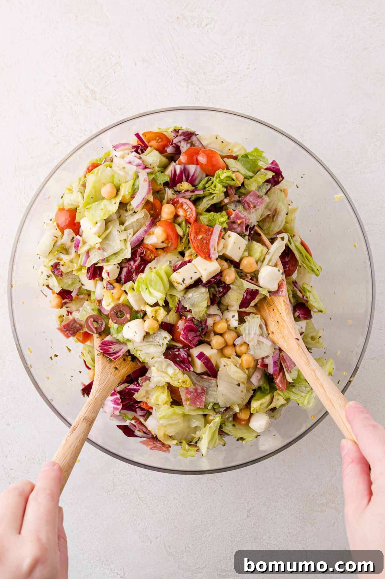Chopped Salad Perfection 8 A person mixing a salad with wooden utensils in a glass bowl, containing lettuce, radicchio, chickpeas, mozzarella, olives, and cherry tomatoes.