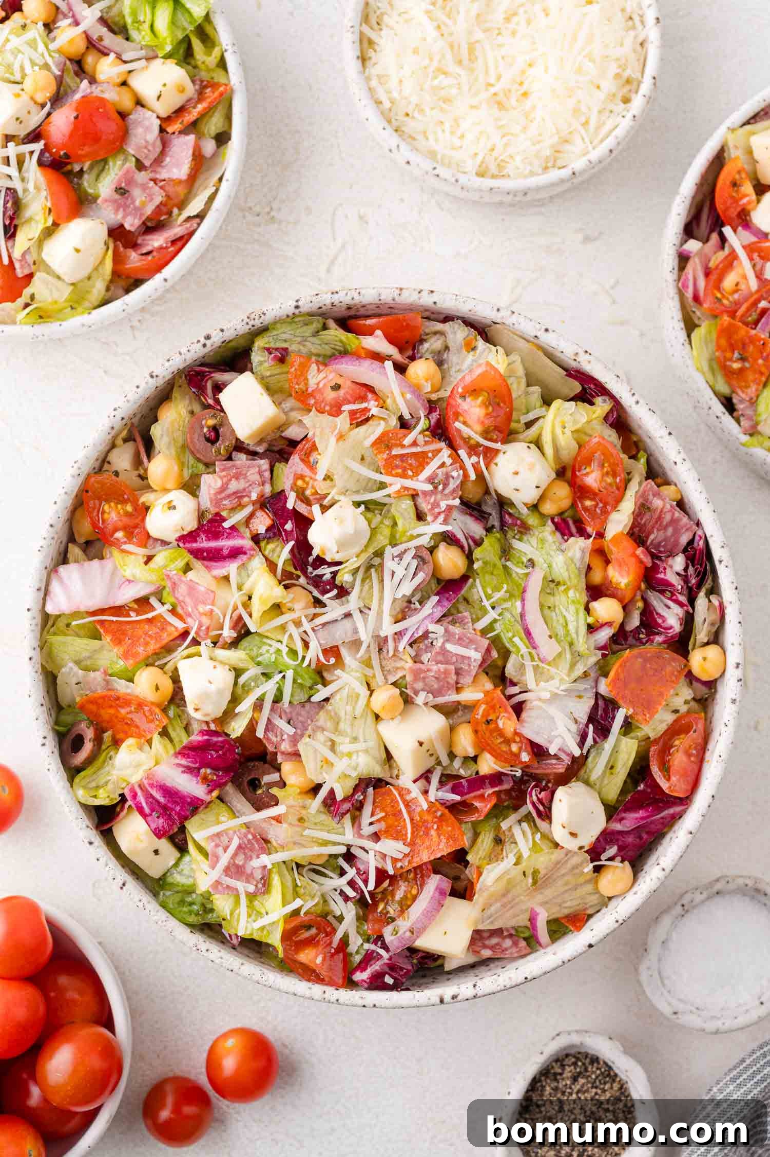 Chopped Salad Perfection 9 A bowl of Italian chopped salad with lettuce, tomatoes, cheese, chickpeas, and cured meats, surrounded by small bowls of grated cheese, cherry tomatoes, and pepper.