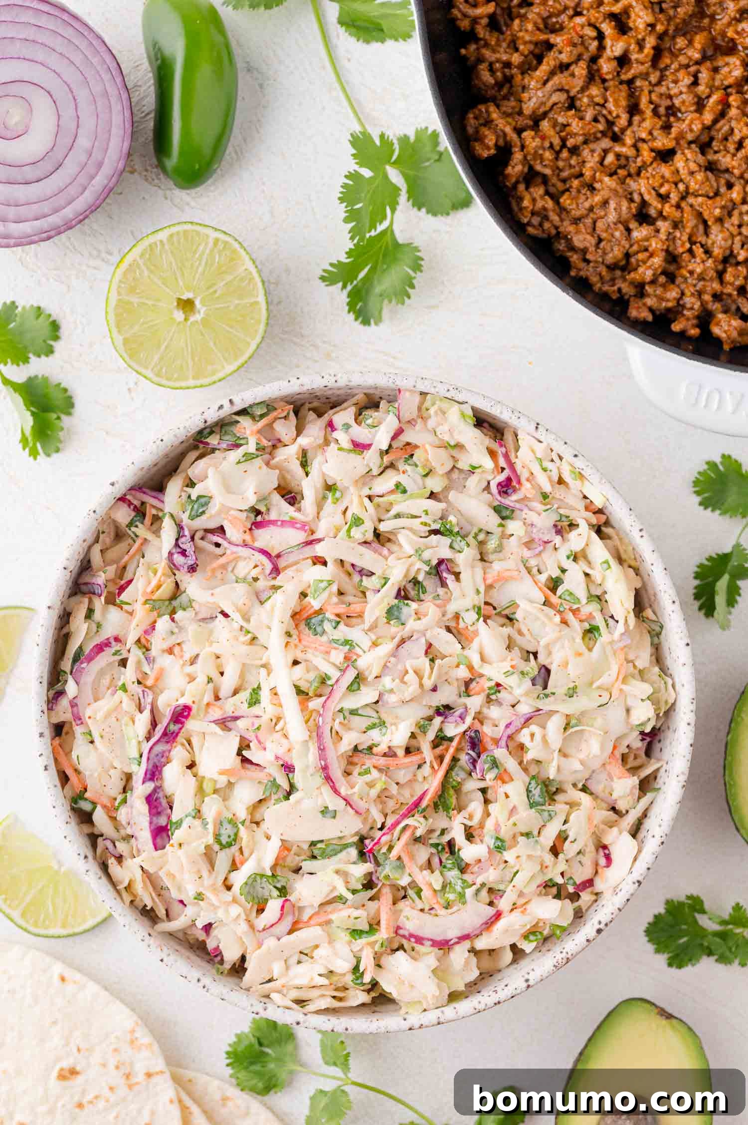 A vibrant bowl of creamy taco slaw featuring shredded cabbage, carrots, finely diced red onions, and fresh cilantro, garnished with lime wedges and surrounded by ingredients like avocado, jalapeño, ground meat, and tortillas on a clean white surface. This slaw adds irresistible crunch, creaminess, and a zippy flavor to your favorite Mexican-inspired dishes.