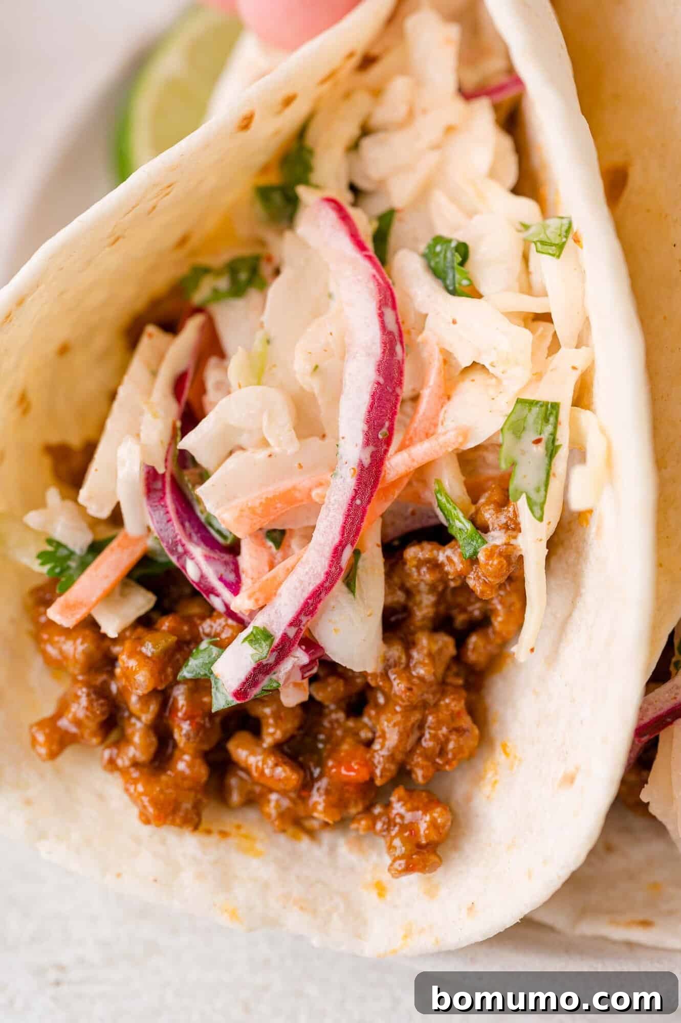 A soft flour taco generously filled with seasoned ground beef and topped with a creamy, colorful slaw made from shredded cabbage, carrots, thin slices of red onion, and fresh green herbs. The vibrant slaw adds a critical crunch and fresh flavor to the savory taco.