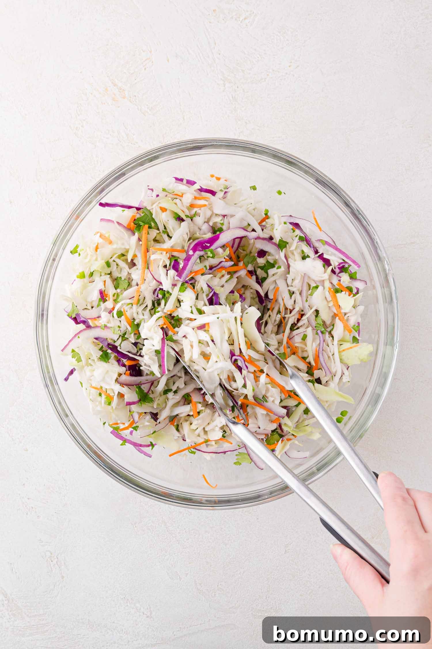 A hand uses metal tongs to gently mix the shredded cabbage, carrots, red onion, and fresh herbs in a large, clear glass bowl on a light-colored surface. The mixing ensures all dry ingredients are well combined before the dressing is added.
