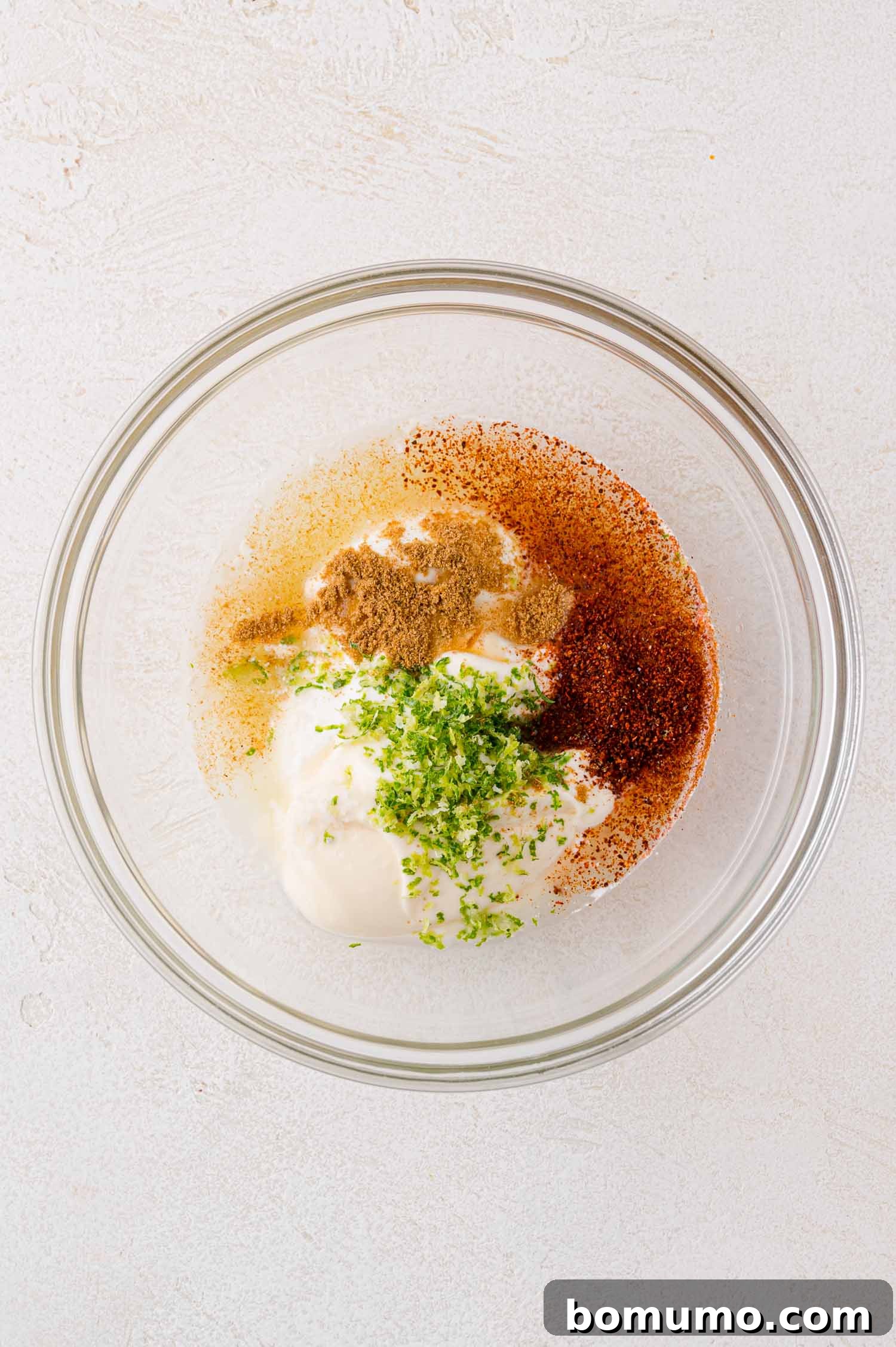 A small glass bowl holding a white, creamy base, soon to be transformed into dressing. It is topped with vibrant green herbs, rich brown spices, and reddish seasonings, awaiting a thorough whisking on a light-colored surface.