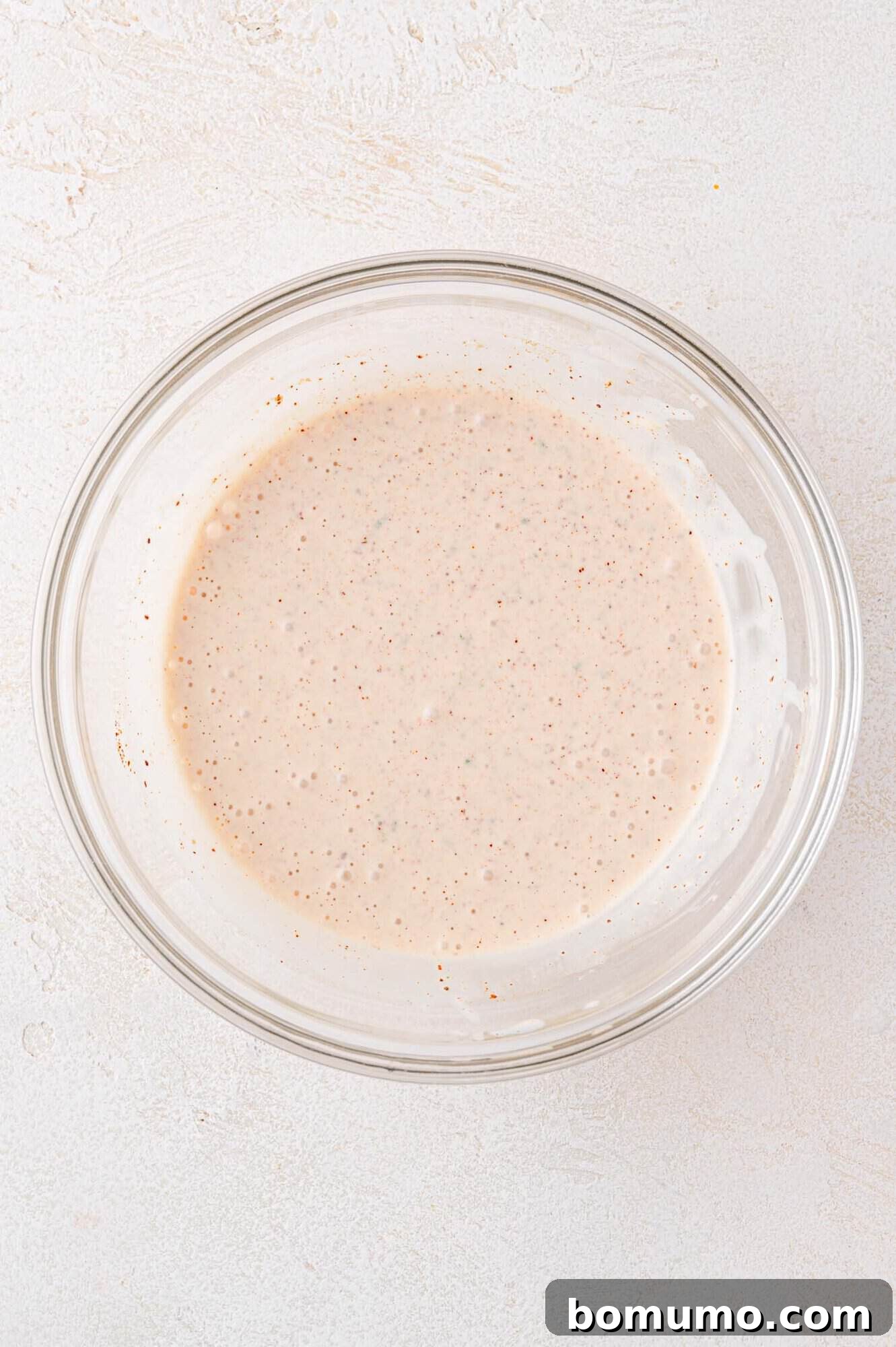 A small glass bowl sits on a light-colored textured surface, holding a creamy, speckled sauce. The dressing, now fully whisked, shows flecks of spices and herbs, ready to be poured over the slaw ingredients.