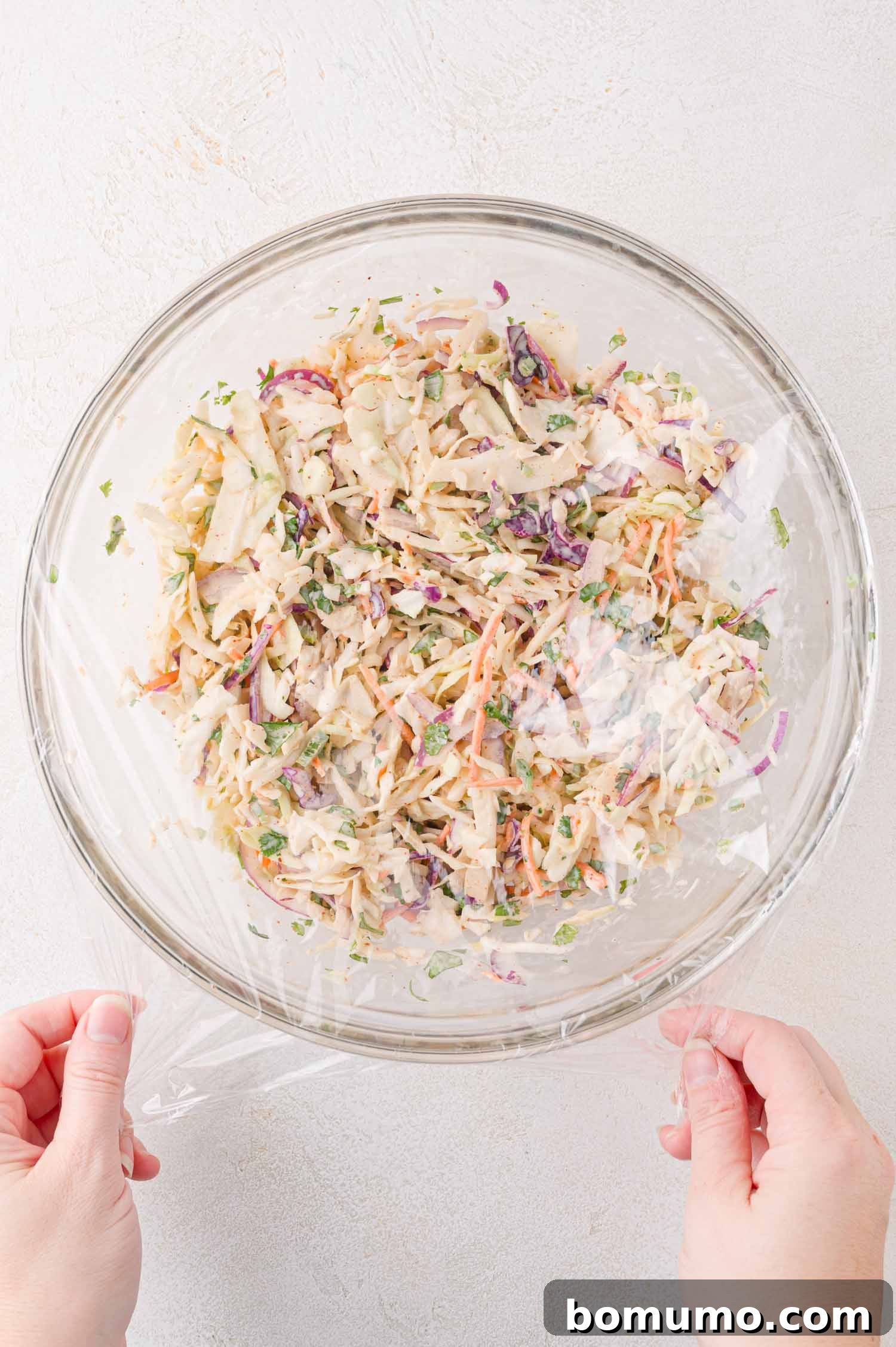 A pair of hands carefully covers a glass bowl filled with freshly prepared coleslaw with a sheet of plastic wrap on a light-colored surface. This step is crucial for allowing the flavors to meld during refrigeration.