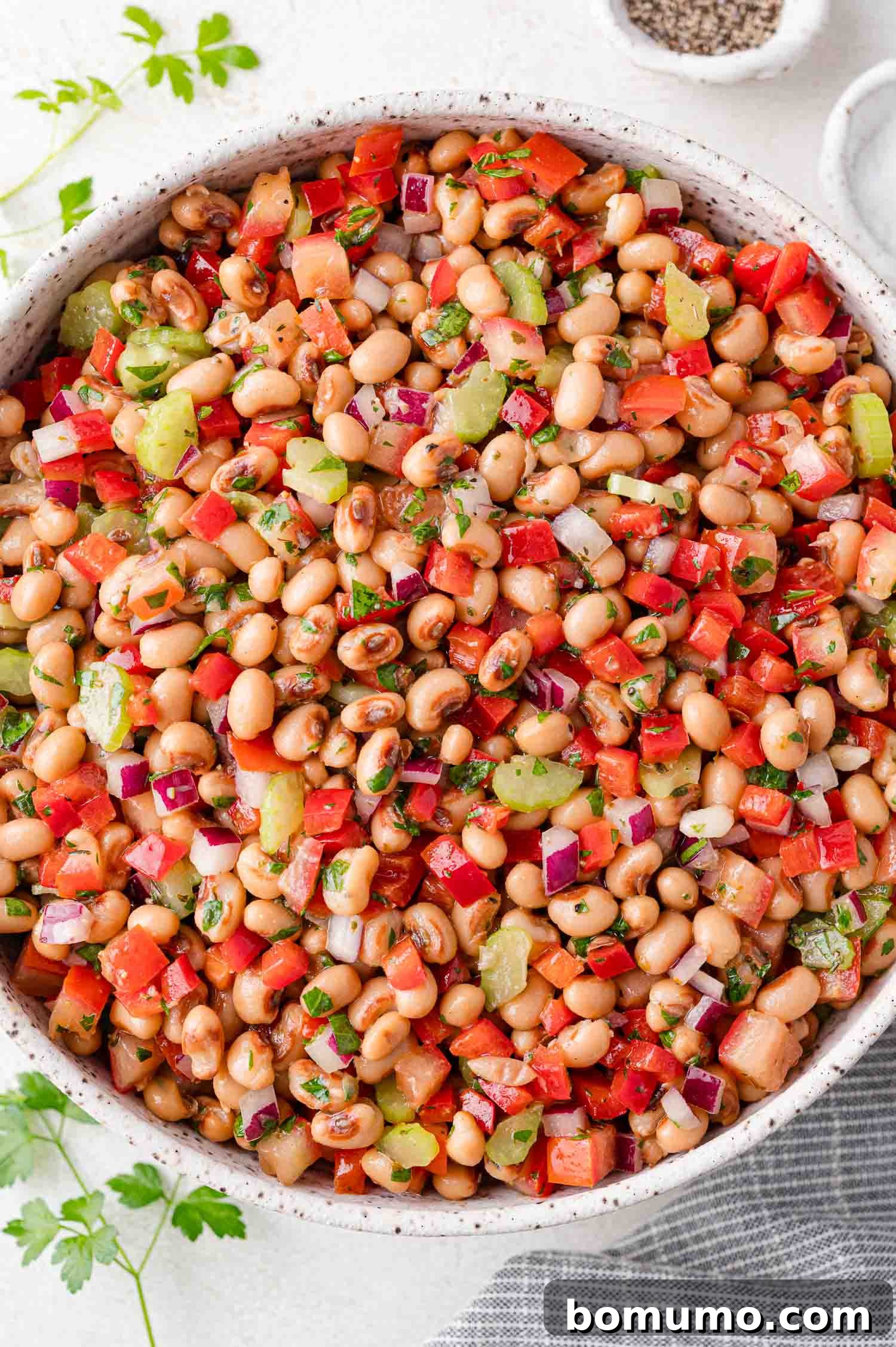 A vibrant bowl of black-eyed pea salad with diced red peppers, ripe tomatoes, crisp celery, red onions, and fresh herbs, served on a light surface with parsley garnish nearby.