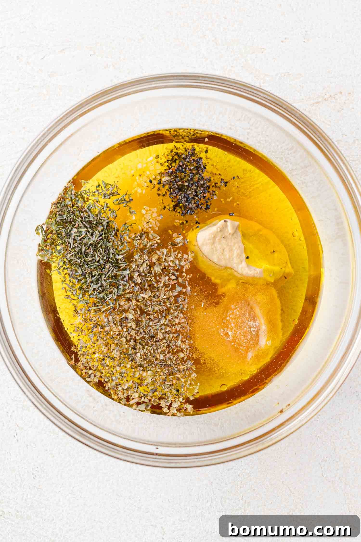 A clear glass bowl holding the liquid ingredients for the salad dressing, including olive oil, vinegar, and mustard, on a pristine white surface.