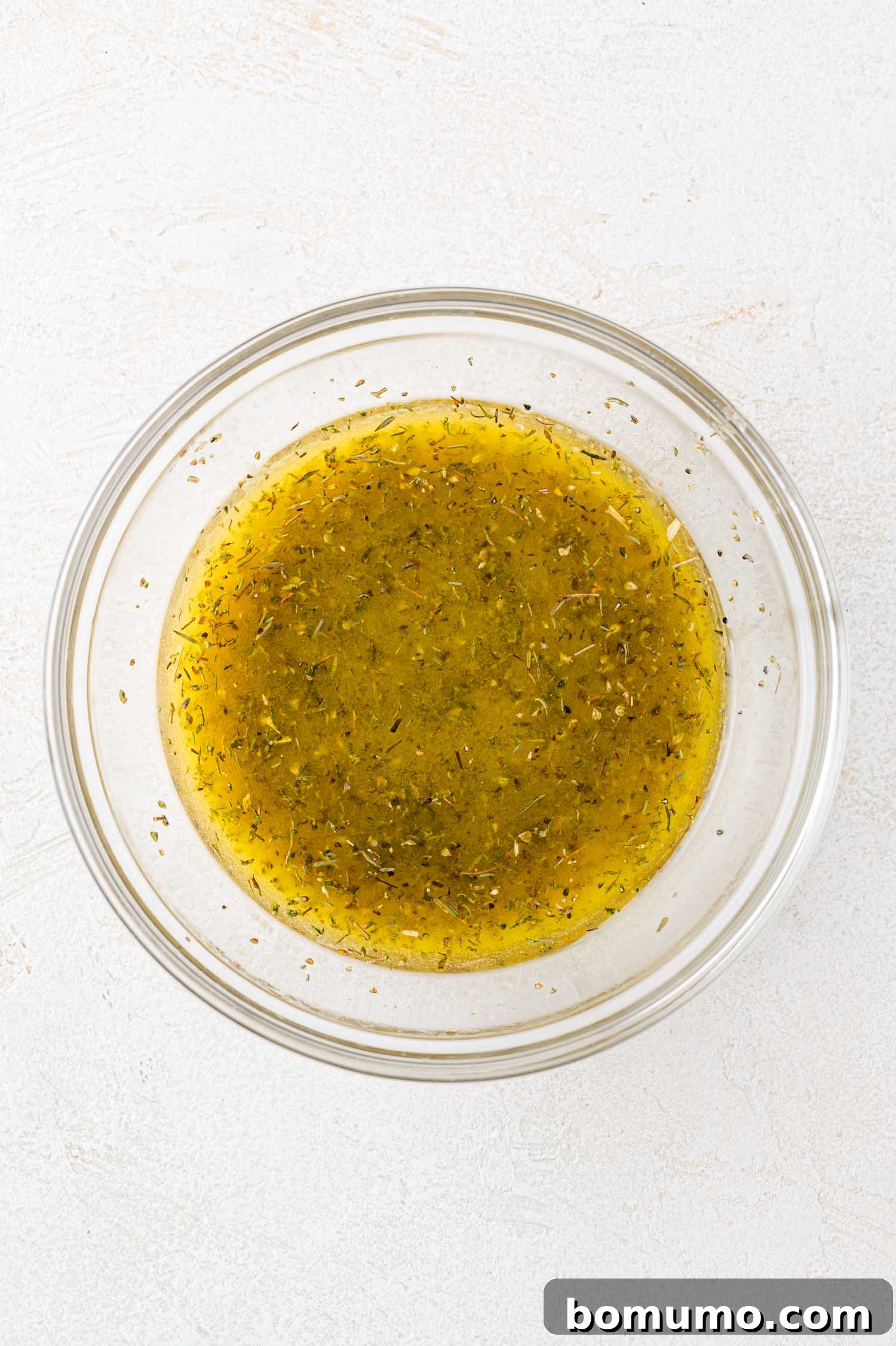 A clear glass bowl filled with a creamy, yellowish vinaigrette dressing, showing subtle flecks of herbs, set on a bright, light-colored surface.