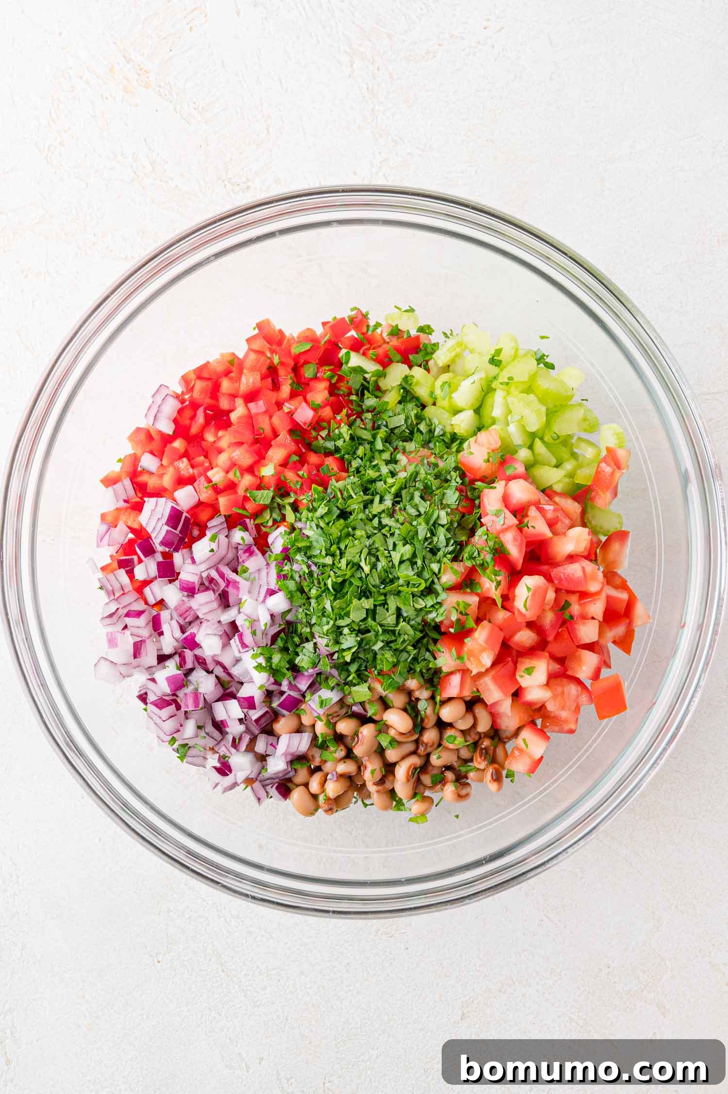 A clear glass bowl showcasing an arrangement of colorful, pre-prepped ingredients: black-eyed peas, diced red onions, tomatoes, celery, red bell pepper, and chopped parsley, ready for mixing.