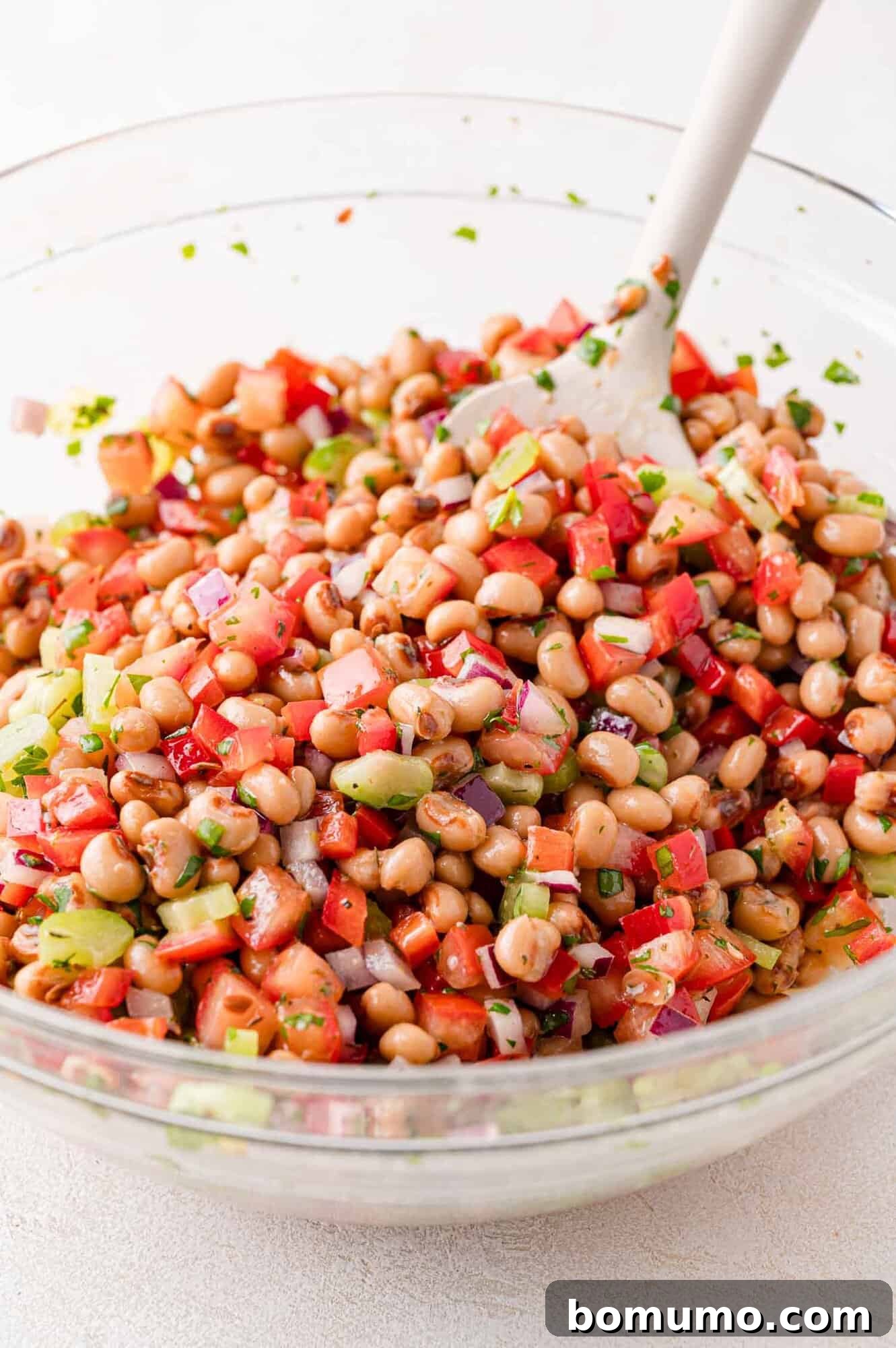 A clear glass bowl filled with a vibrant mix of black-eyed pea salad ingredients, including peas, diced red tomatoes, red onion, celery, and fresh herbs, with a white mixing spoon inside.