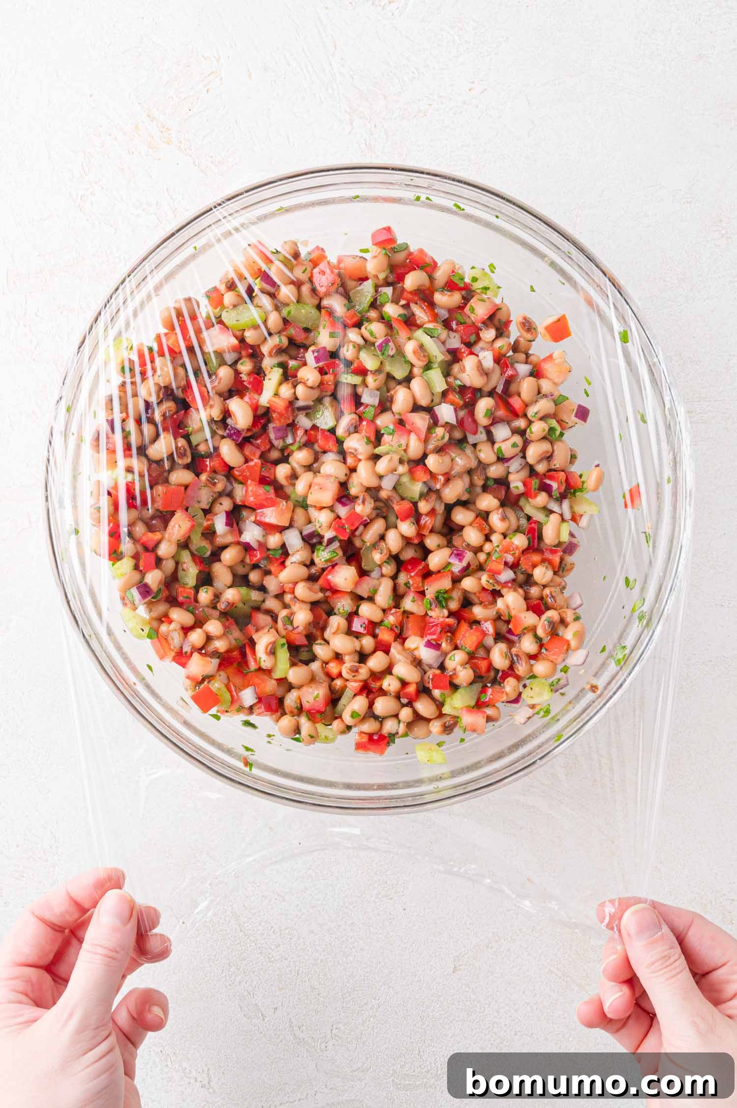 A person covers a glass bowl of freshly mixed black-eyed pea salad with clear plastic wrap on a clean white surface, preparing it for refrigeration.