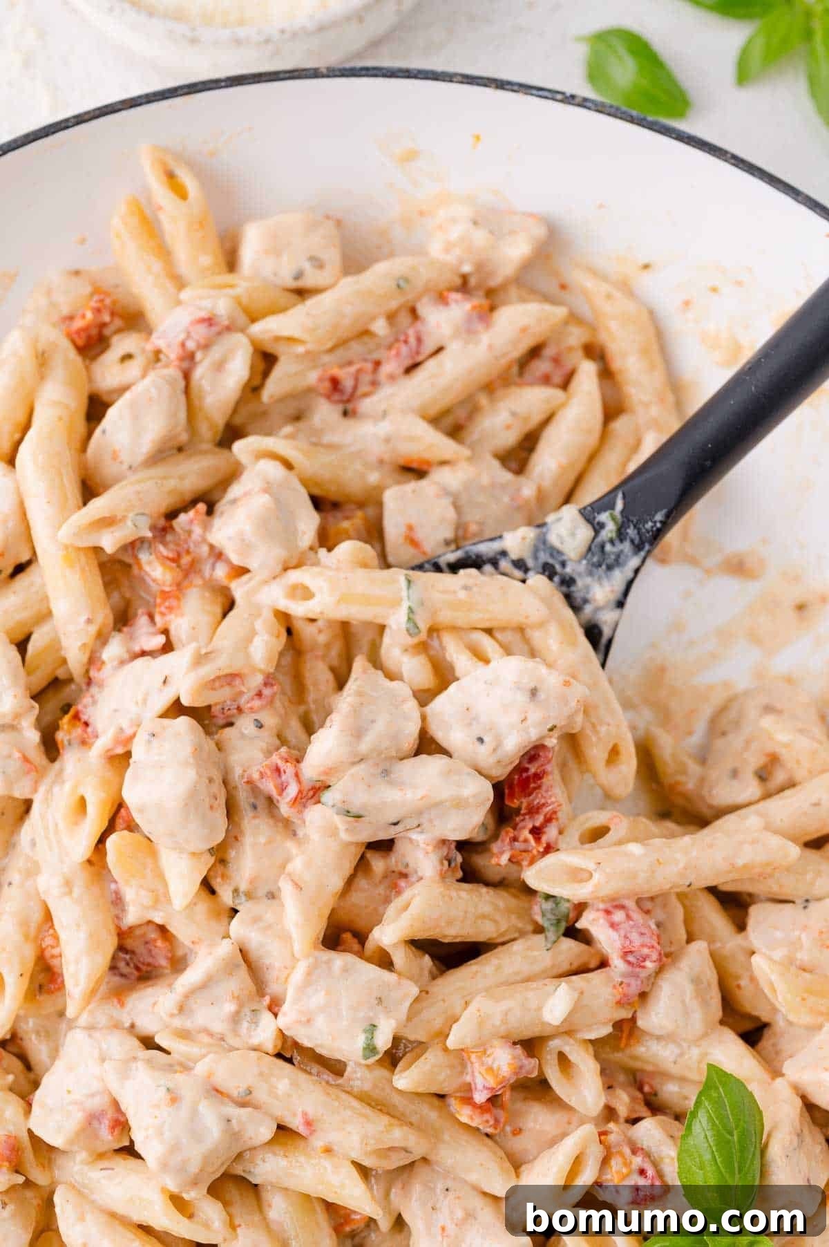 Close-up of creamy pasta with penne, chunks of chicken, and vibrant sun-dried tomatoes in a skillet. Garnished generously with fresh basil leaves.
