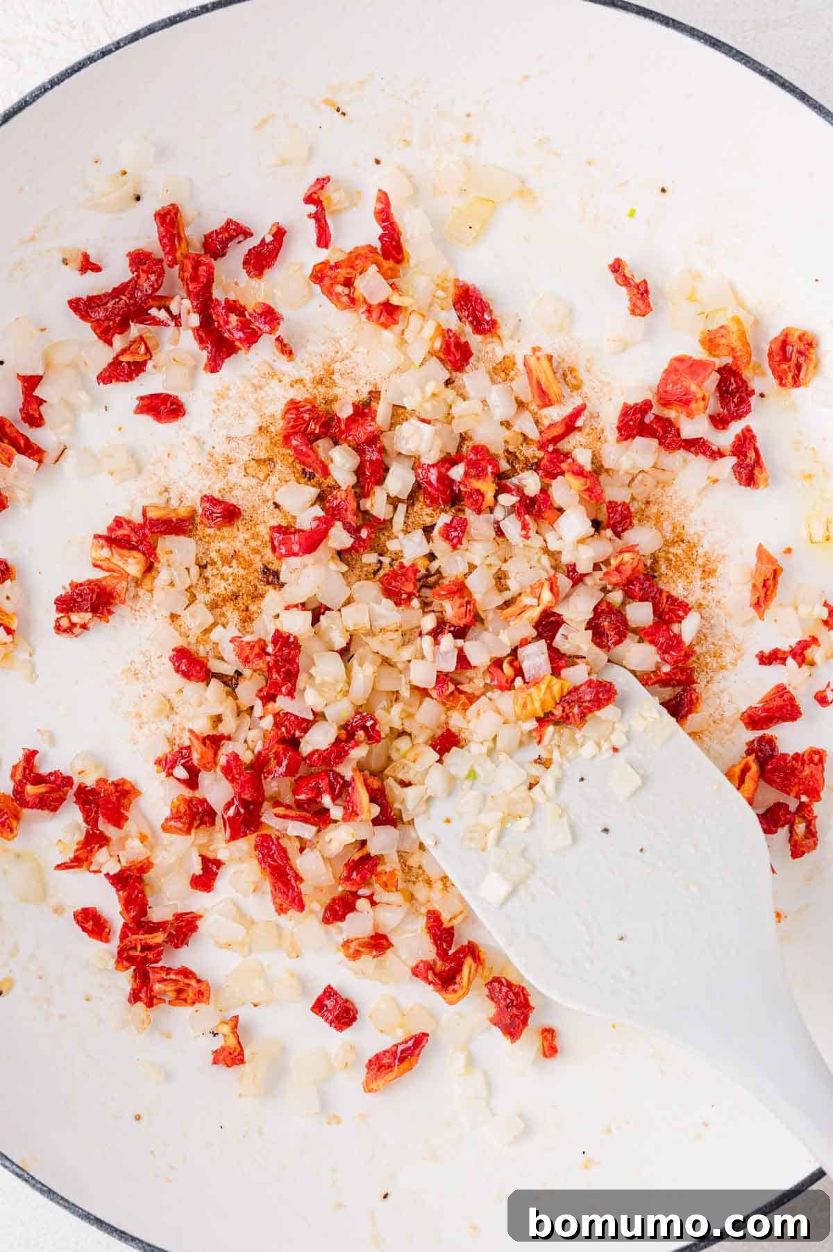 A mix of diced onions and sun-dried tomatoes being sautéed in a white pan, stirred with a white spatula, releasing their enticing aromas.