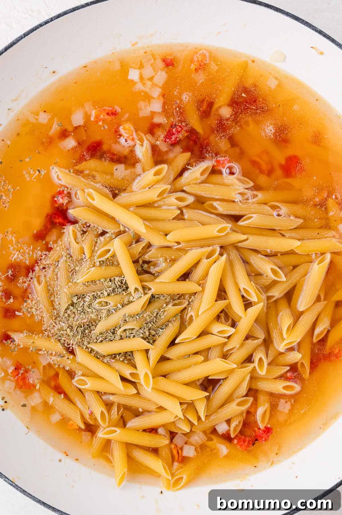 A pot filled with uncooked penne pasta, vibrant sun-dried tomatoes, finely chopped onions, and fragrant herbs submerged in a savory liquid base, ready to simmer.
