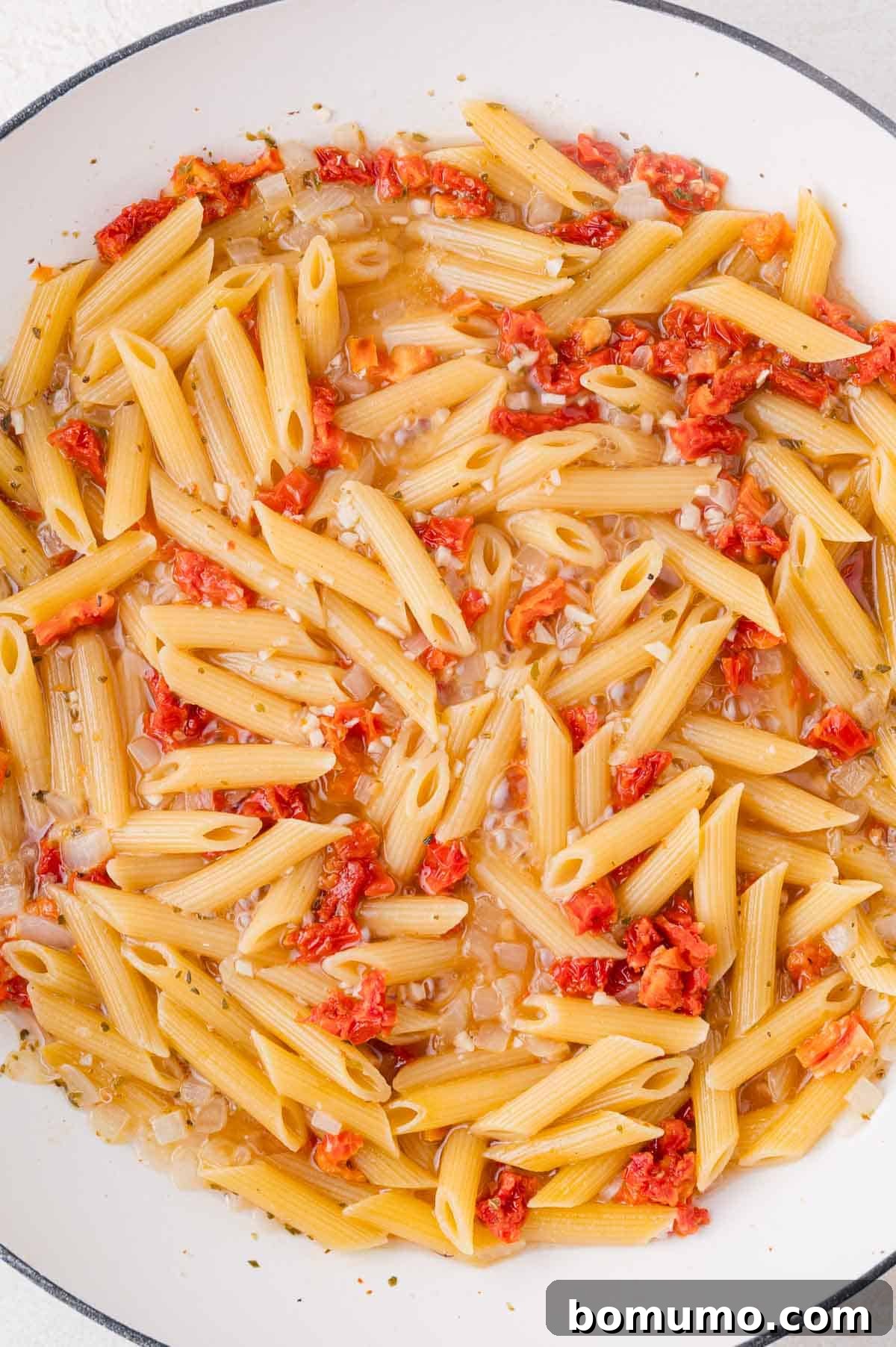A white pan containing perfectly cooked penne pasta, infused with diced sun-dried tomatoes and finely chopped onions, showing absorption of liquid.