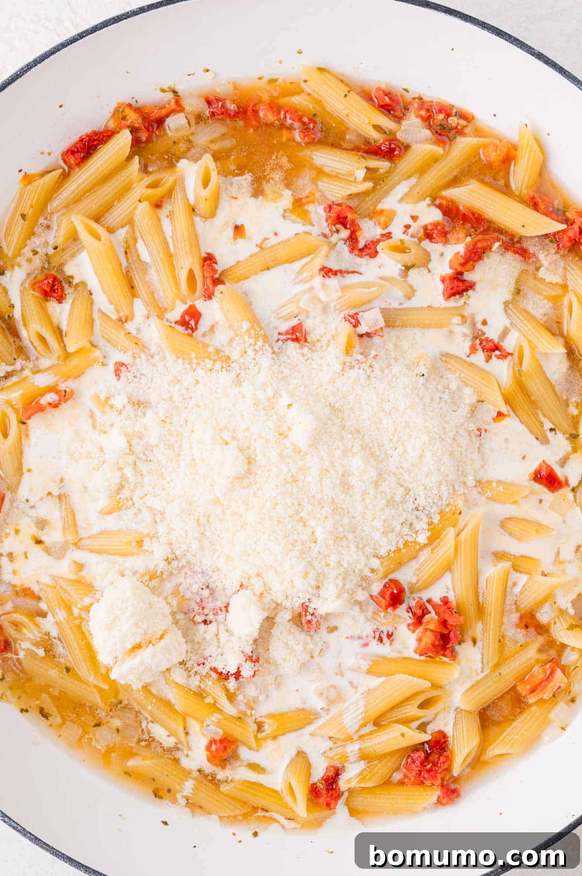 A white pan showcasing perfectly cooked penne pasta enveloped in a rich cream sauce, studded with sun-dried tomatoes, and topped with a generous pile of grated cheese, ready to be mixed.