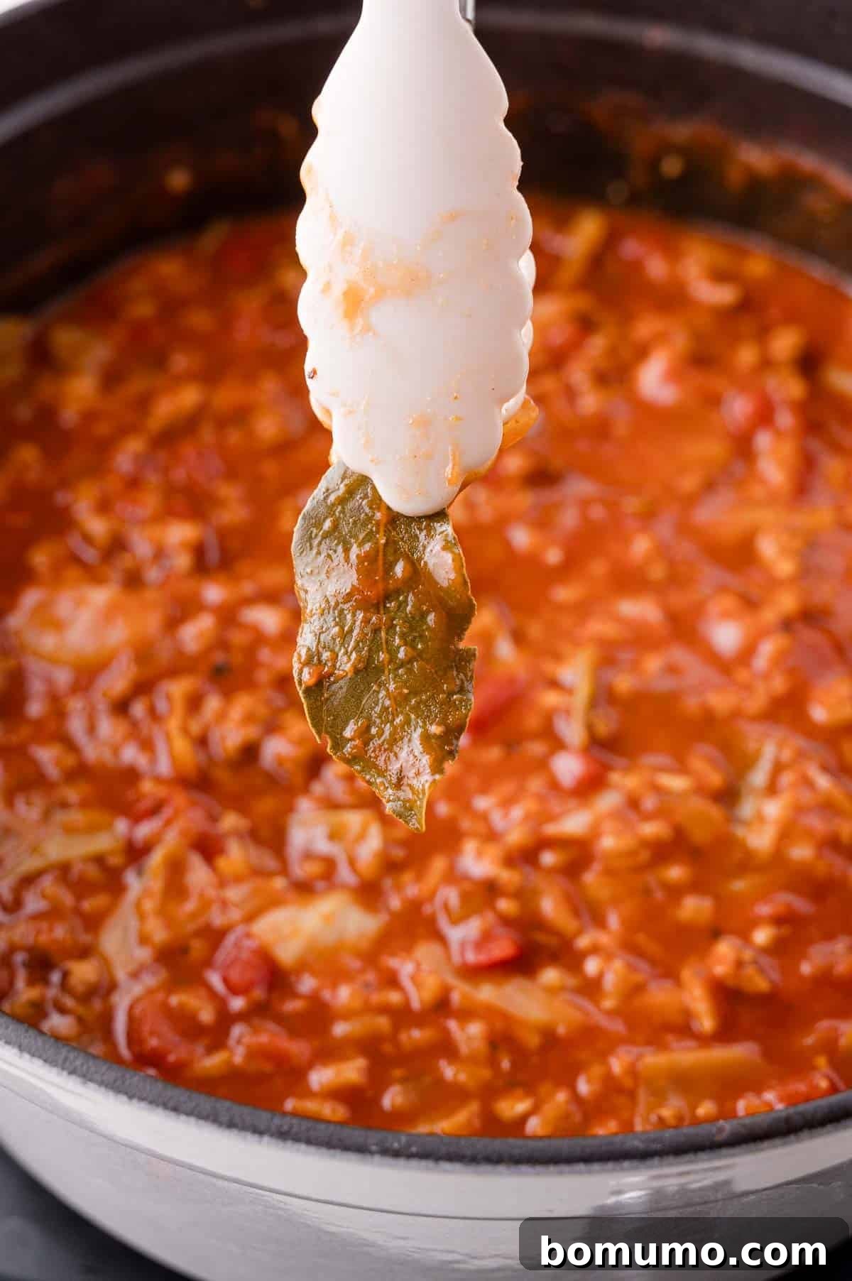 Deconstructed Cabbage Roll Soup 11 A white tong carefully removes a single bay leaf from a steaming pot of rich, red tomato-based soup, which contains visible chunks of meat and vegetables, signaling the soup is nearing completion.
