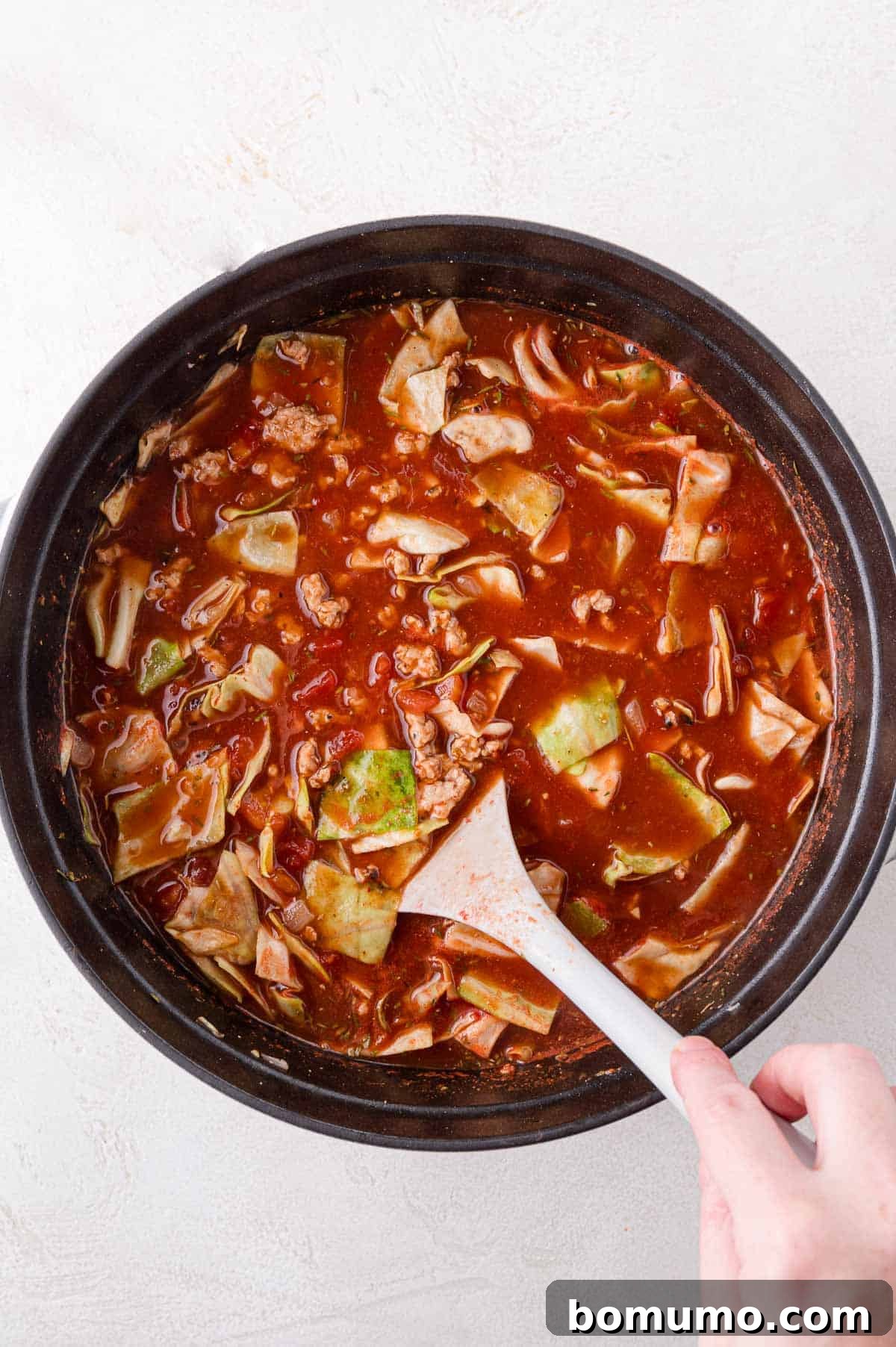 Deconstructed Cabbage Roll Soup 10 A hearty pot of rich, red tomato-based soup gently simmering, showcasing visible chunks of tender cabbage and savory meat. A spatula is actively stirring the contents, ensuring an even distribution of flavors as it cooks.
