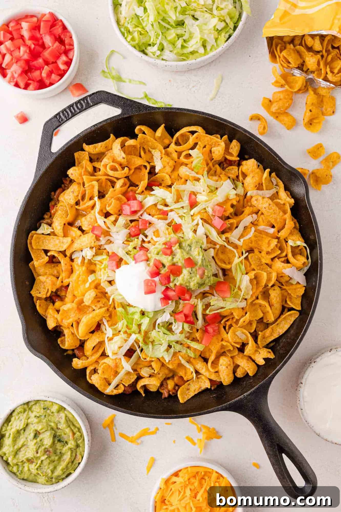 Crunchy Taco Casserole 2 Skillet of walking tacos casserole topped with crisp lettuce, fresh tomatoes, and creamy sour cream, surrounded by small bowls of shredded cheese, additional lettuce, diced tomatoes, and rich guacamole.