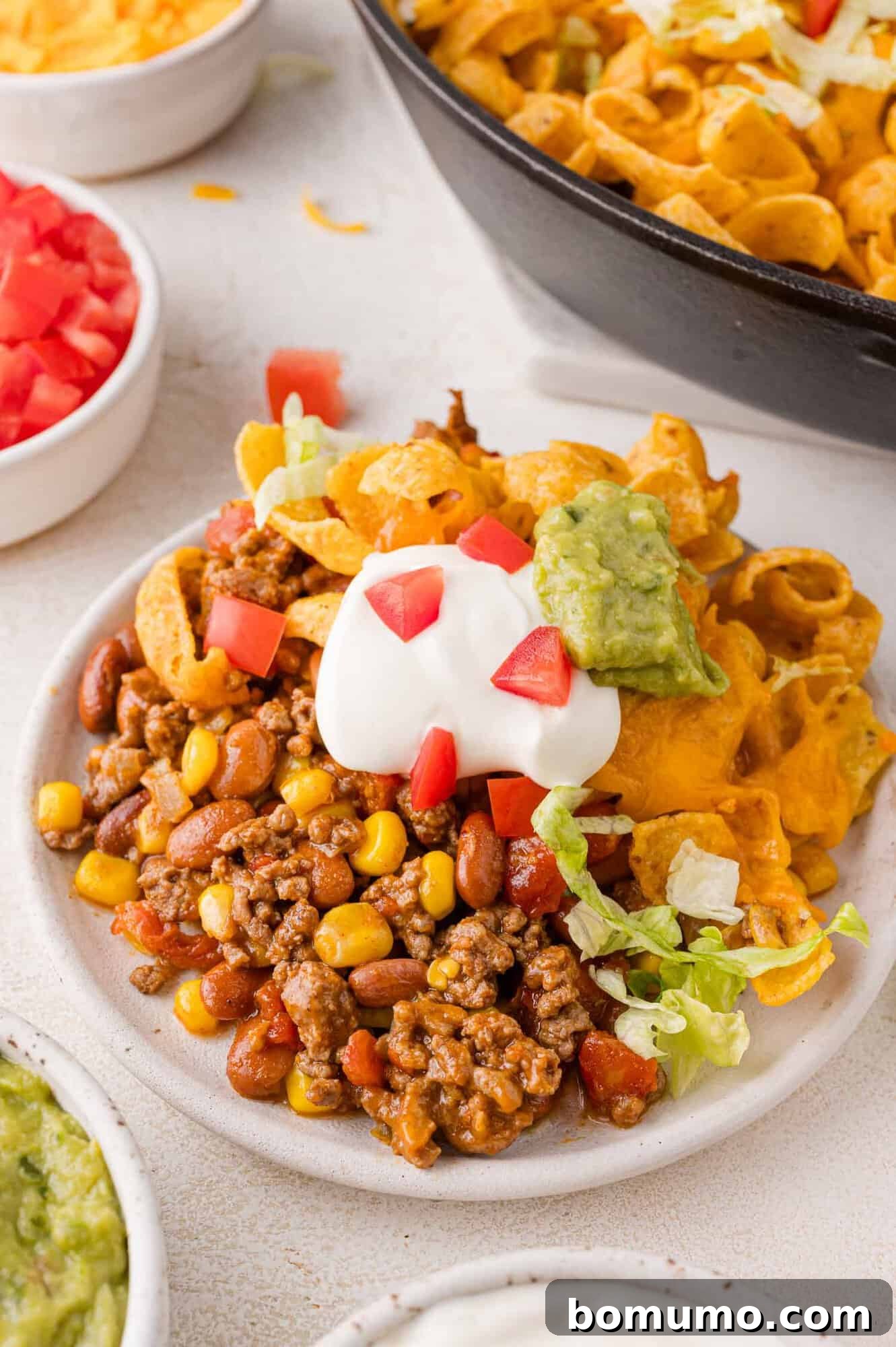 Crunchy Taco Casserole 11 A close-up of a plate piled high with Walking Taco Casserole, showcasing the chili, corn, and beans generously topped with crunchy corn chips, fresh diced tomatoes, creamy sour cream, rich guacamole, and crisp shredded lettuce. Nearby bowls offer additional ingredients for customization, inviting diners to create their perfect bite.