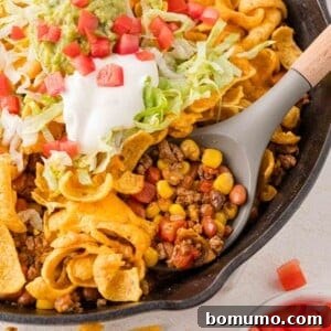 A skillet filled with taco ingredients, including ground beef, corn, beans, and topped with sour cream, guacamole, shredded lettuce, tomatoes, and corn chips, showcasing a complete and delicious meal.